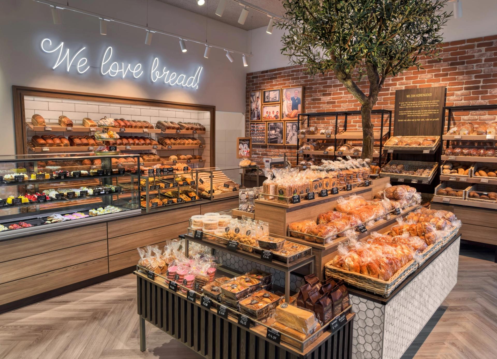 Winkel van Korengoud De Echte Bakker in Rosmalen, met een neonletter We love bread, een olijfboom en broodmeubels op een visgraatvloer.
