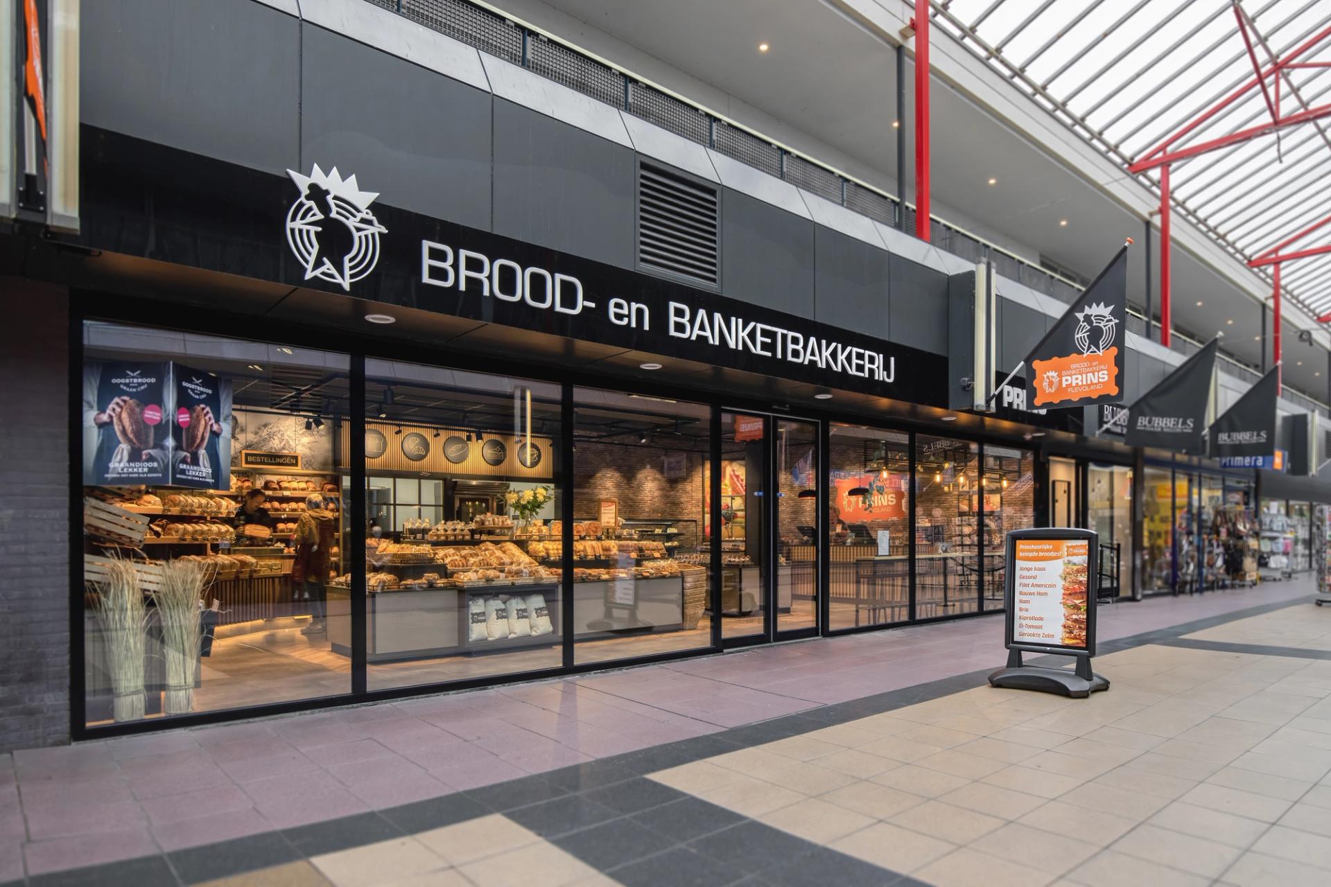 “Entree en winkelpui van Brood- en Banketbakkerij Prins in Flevoland, met een moderne zwarte gevel en zicht op de sfeervolle bakkerij en uitgebreide broodpresentatie.”