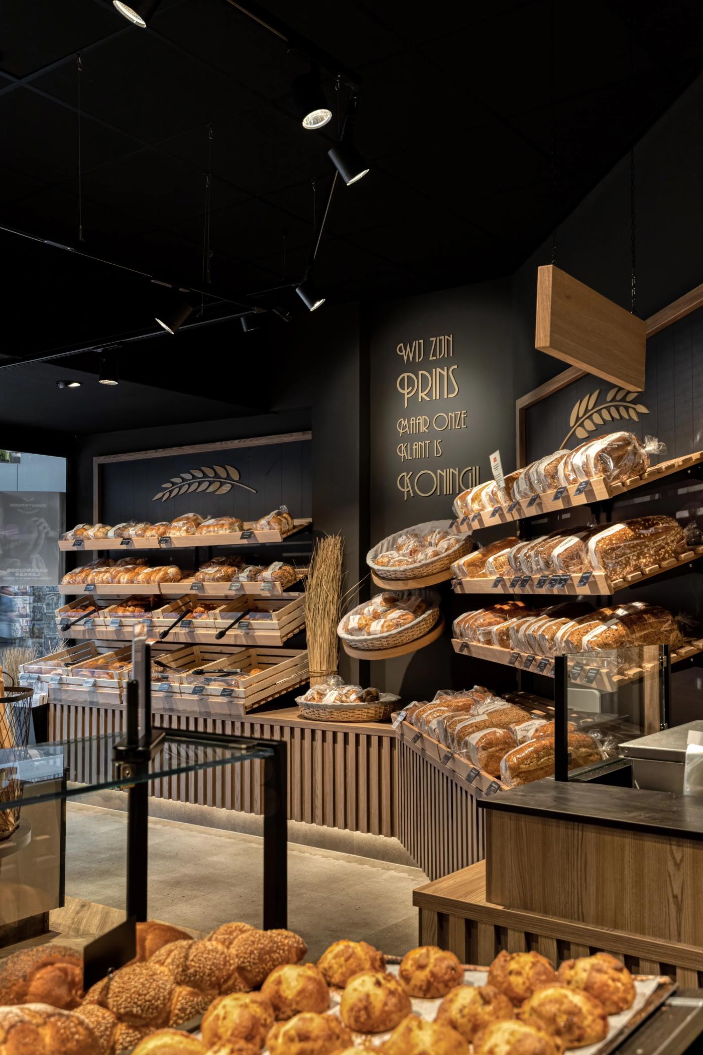 “Ambachtelijke broodpresentatie bij Brood- en Banketbakkerij Prins in Flevoland, met houten broodrekken, warme verlichting en een stoer zwart interieur met de tekst ‘Wij zijn Prins, maar onze klant is koning’.”