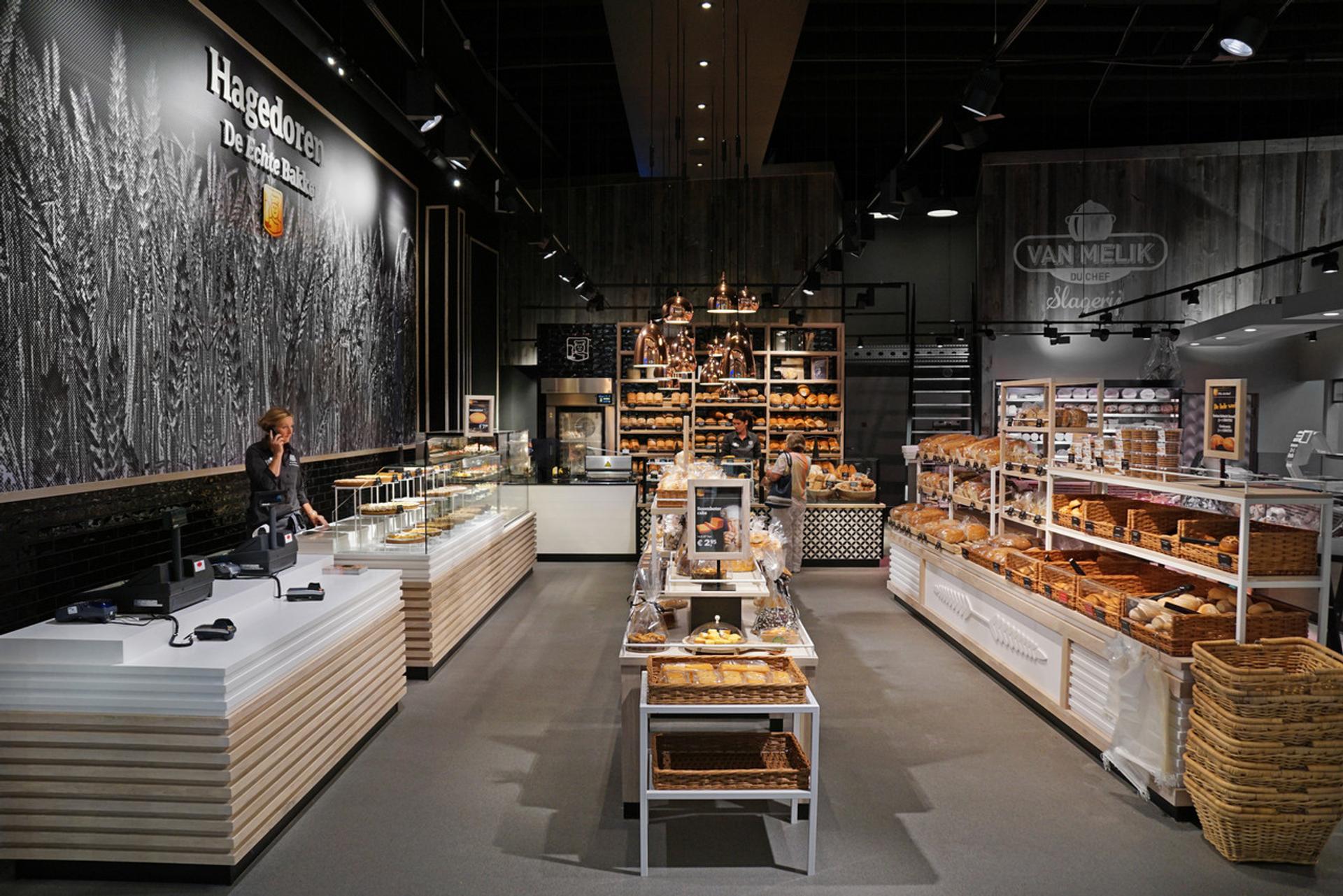 Interieur van Echte Bakker Hagedoren in Beek, Limburg, met moderne brood- en gebakspresentaties, warme houtaccenten, industriële verlichting en een grote wandvisual die het ambachtelijke karakter van de bakkerij benadrukt.