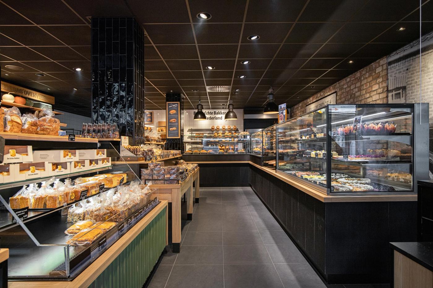 Sfeervol winkelinterieur van Bakkerij Gommans in Horst met royale brood- en gebakspresentaties, warme houtaccenten, groene fronten en moderne zwarte vitrines.
