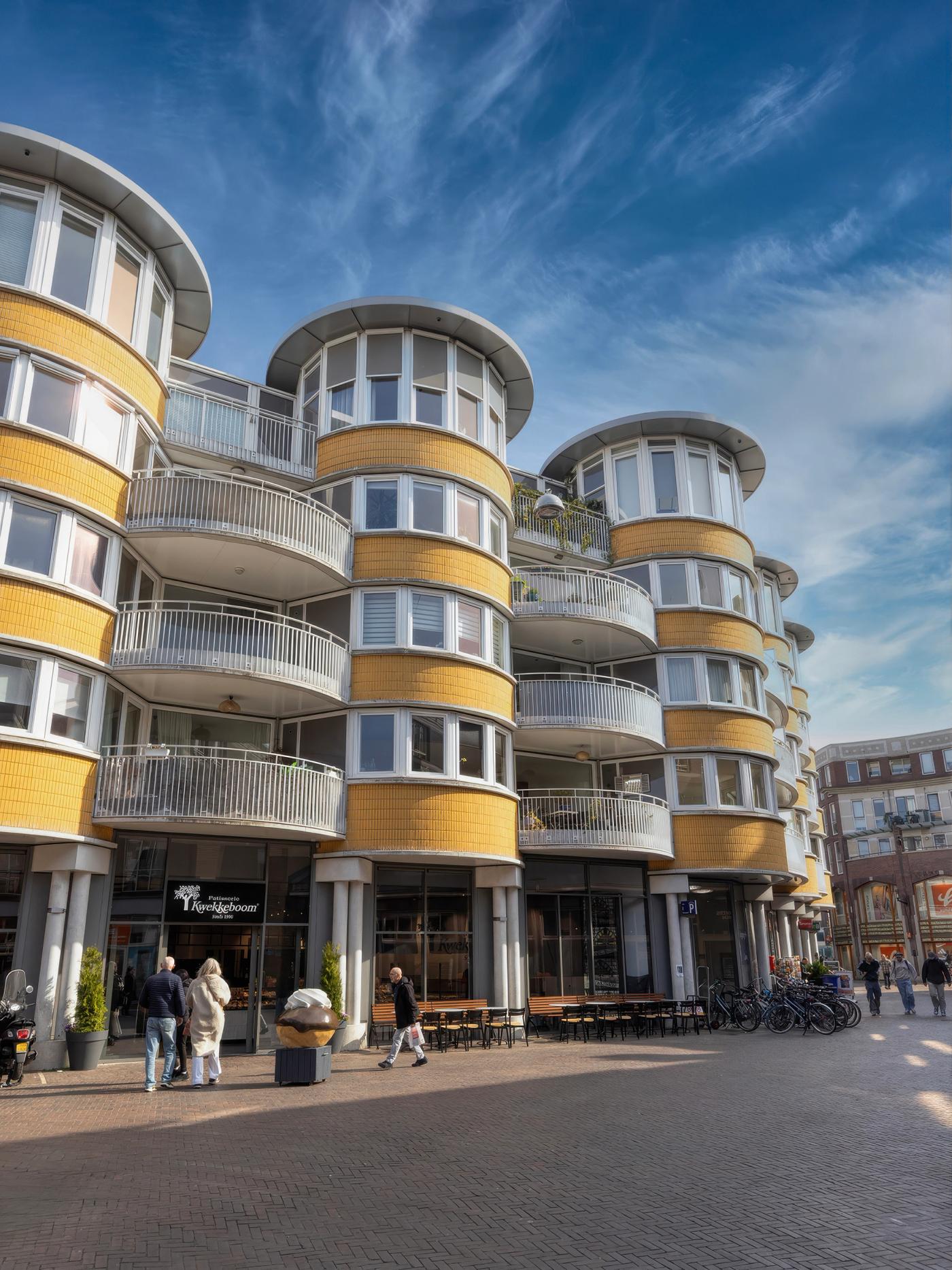 “Patisserie Kwekkeboom in Amsterdam, gevestigd op de begane grond van een markant stedelijk pand met ronde gevels en terrassen, met winkel en zitgelegenheid aan het Land van Cocagneplein.”