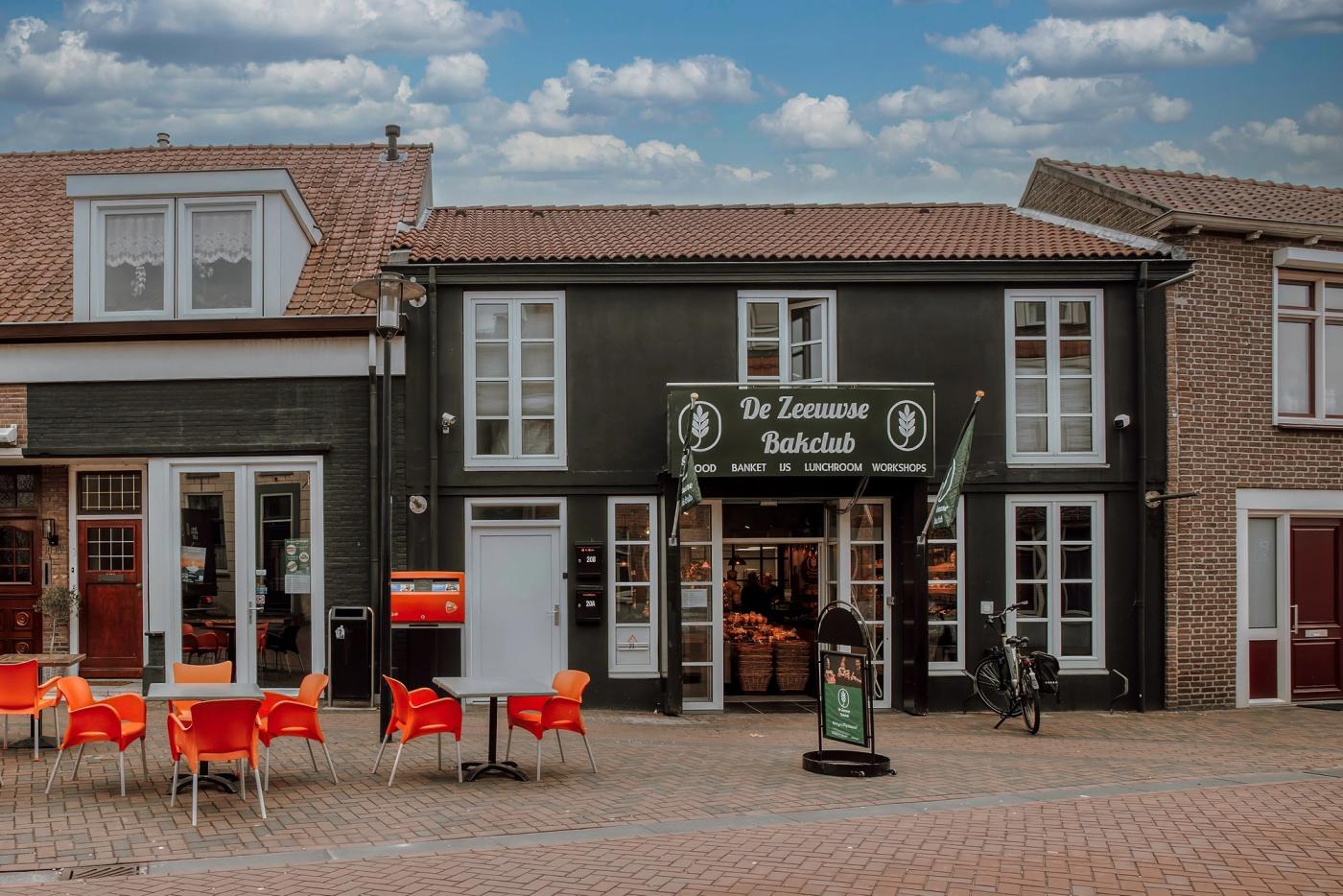 Gevel van De Zeeuwse Bakclub in Kruiningen, met een terras met oranje stoelen voor de entree.