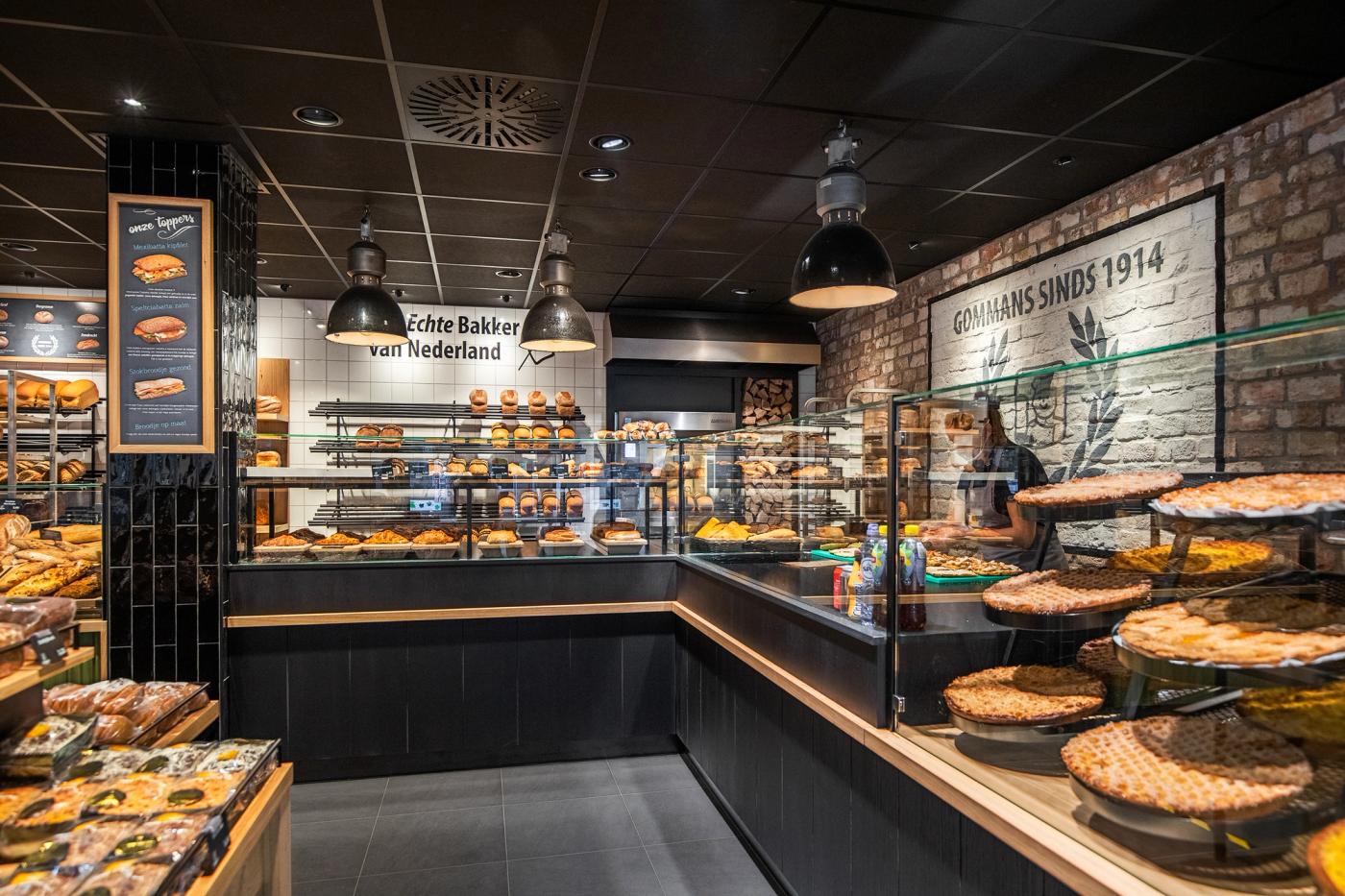 Sfeervol winkelinterieur van Bakkerij Gommans in Horst met royale brood- en gebakspresentaties, warme houtaccenten, groene fronten en moderne zwarte vitrines.