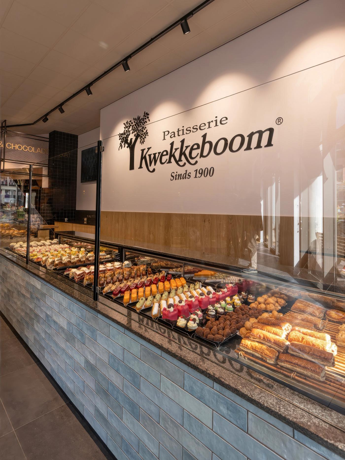 “Luxe gebakspresentatie bij Patisserie Kwekkeboom in Amsterdam met kleurrijk patisserie-assortiment, blauw betegelde toonbank en het karakteristieke Kwekkeboom-logo.”