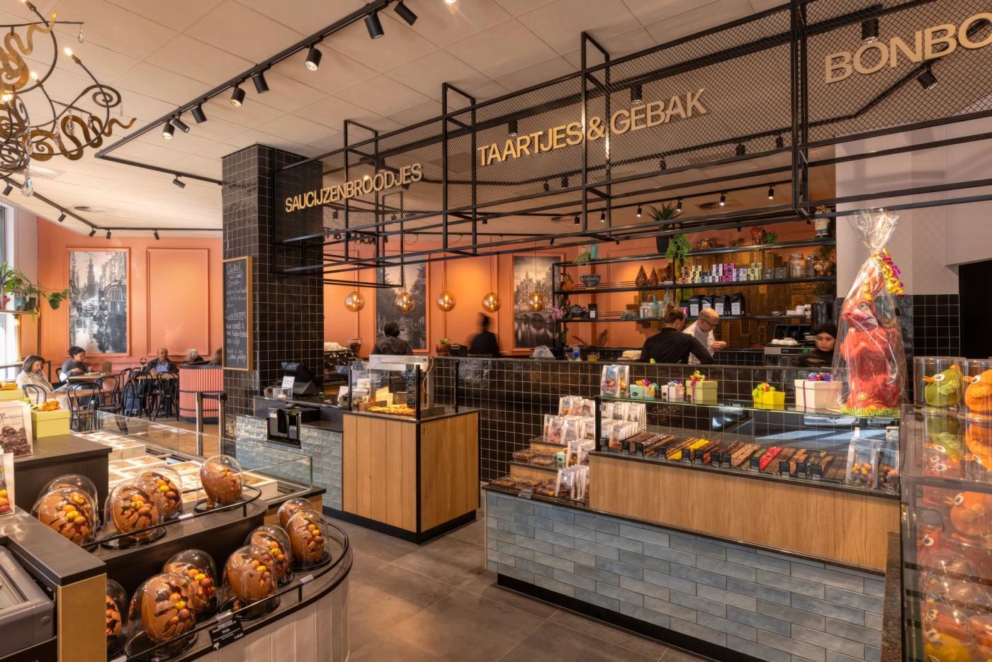 “Interieur van Patisserie Kwekkeboom in Amsterdam met luxe chocoladepresentatie, gebak en saucijzenbroodjes, warme roze tinten, hout, zwarte tegels en industriële details.”