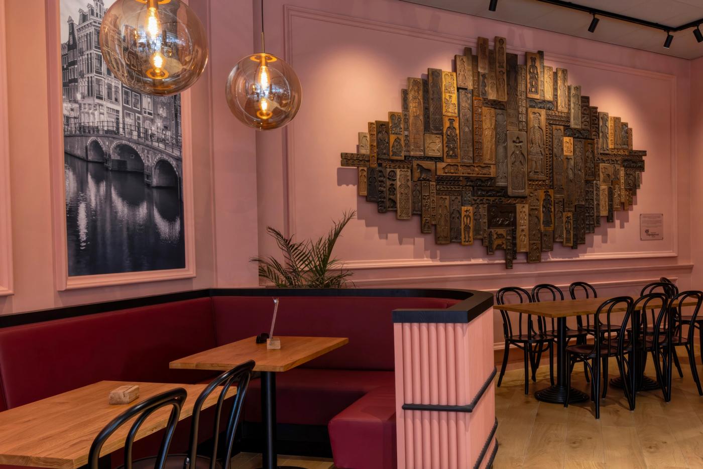“Historische speculaasplanken als kunstwerk aan de wand in de sfeervolle lunchroom van Patisserie Kwekkeboom in Amsterdam, gecombineerd met warme roze tinten, houten tafels en klassieke verlichting.”