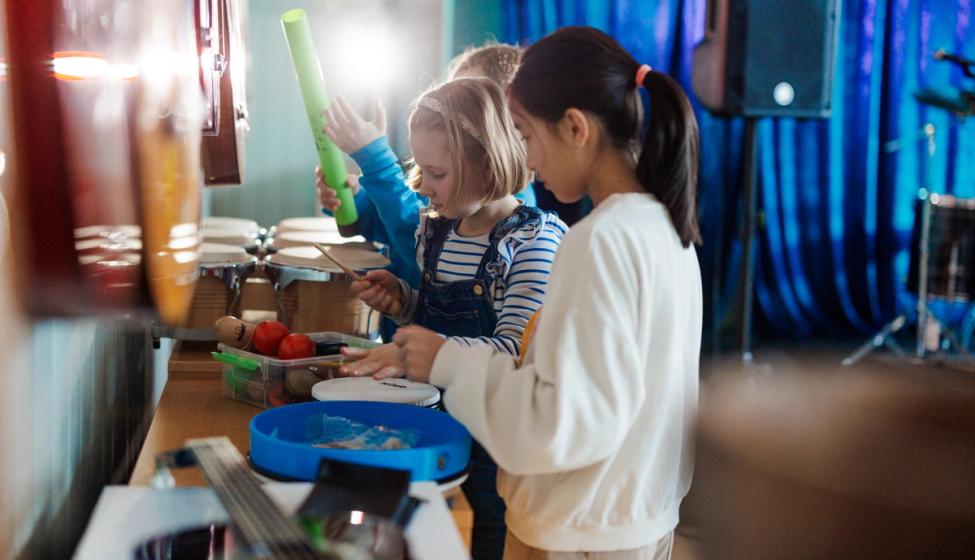 Barn tester ut ulike rytmeinstrumenter i et musikkrom. På veggen henger gitarer, i bakgrunnen ser vi en høyttaler og et sceneteppe.