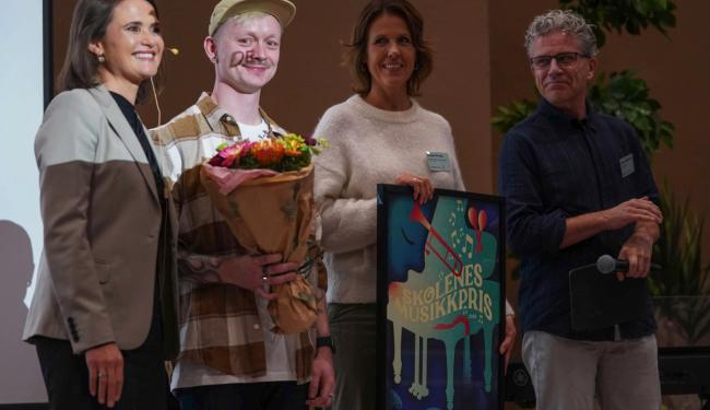 Fire personer på scenen - vinnerne i midten holder blomster og en innrammet plakat.