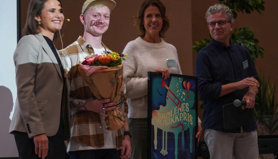 Fire personer på scenen - vinnerne i midten holder blomster og en innrammet plakat.
