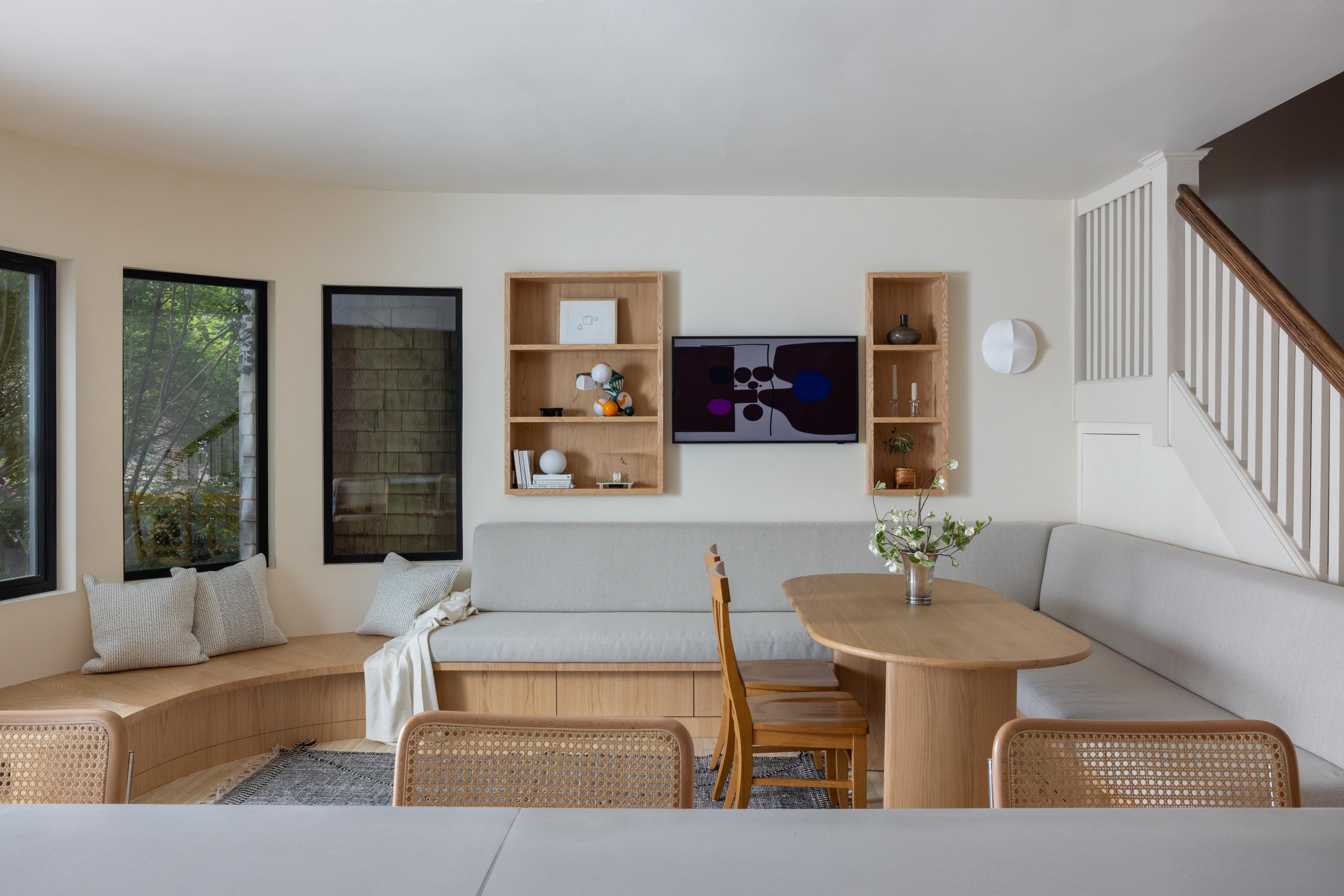 Dining room with curved banquette, seat upholstered in light gray, light-wood dining table. Two floating oak bookcases with TV in the middle.