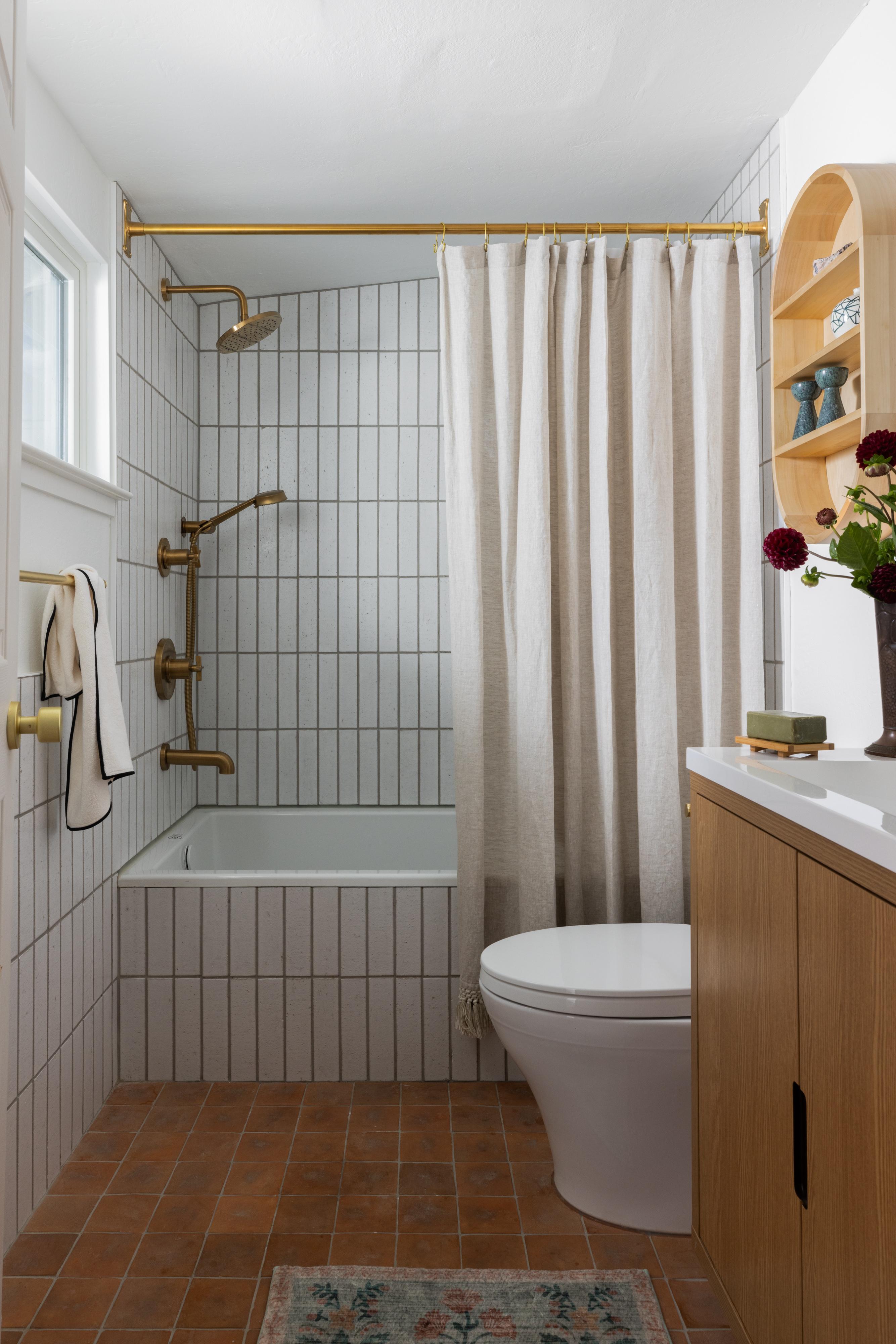 Carmichael bathroom with a white tiled tub and shower, brass fixtures, a linen curtain and terracotta floor tile