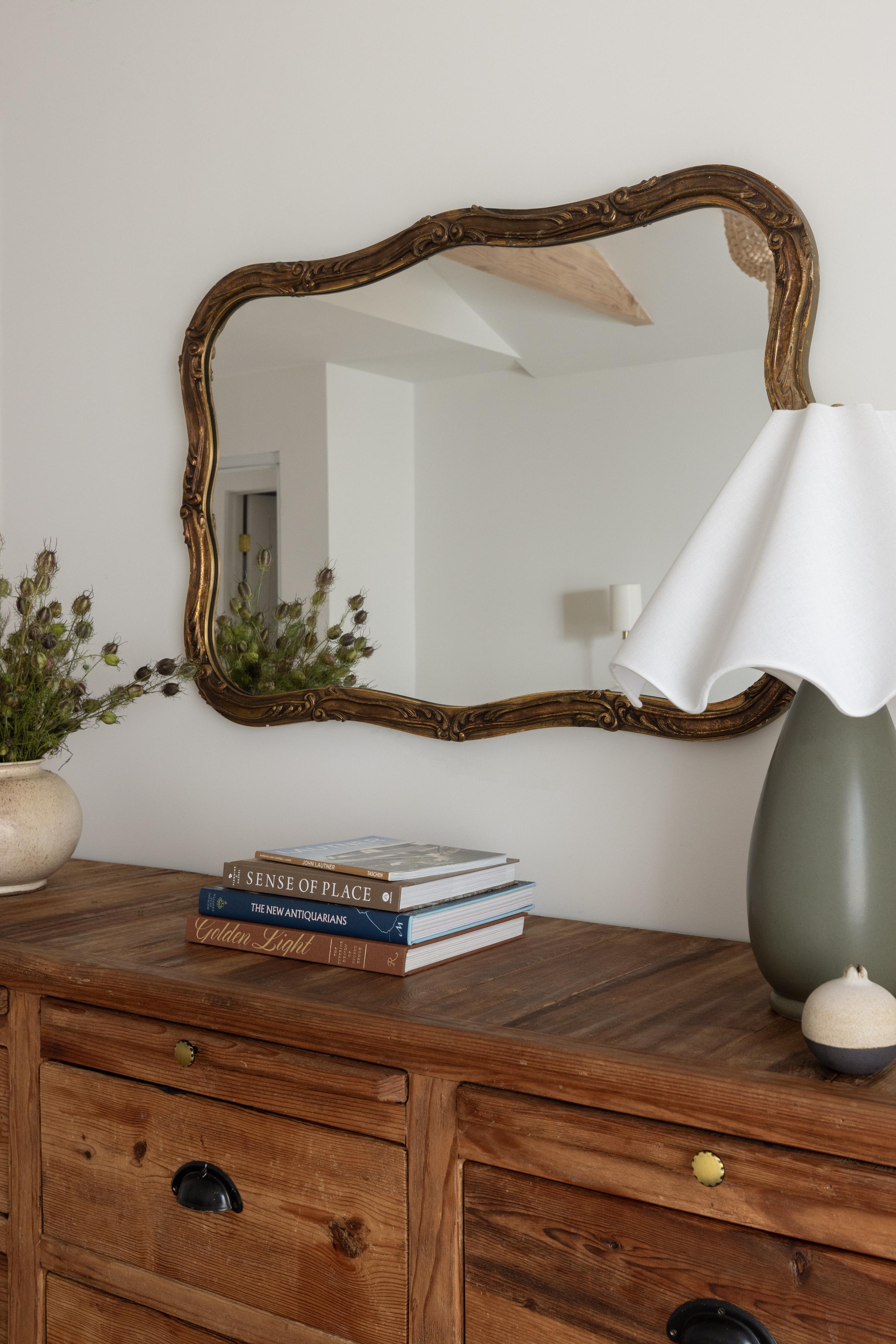Antique gilt mirror above a reclaimed wood dresser with a sculptural lamp and stacked books in a Carmichael bedroom