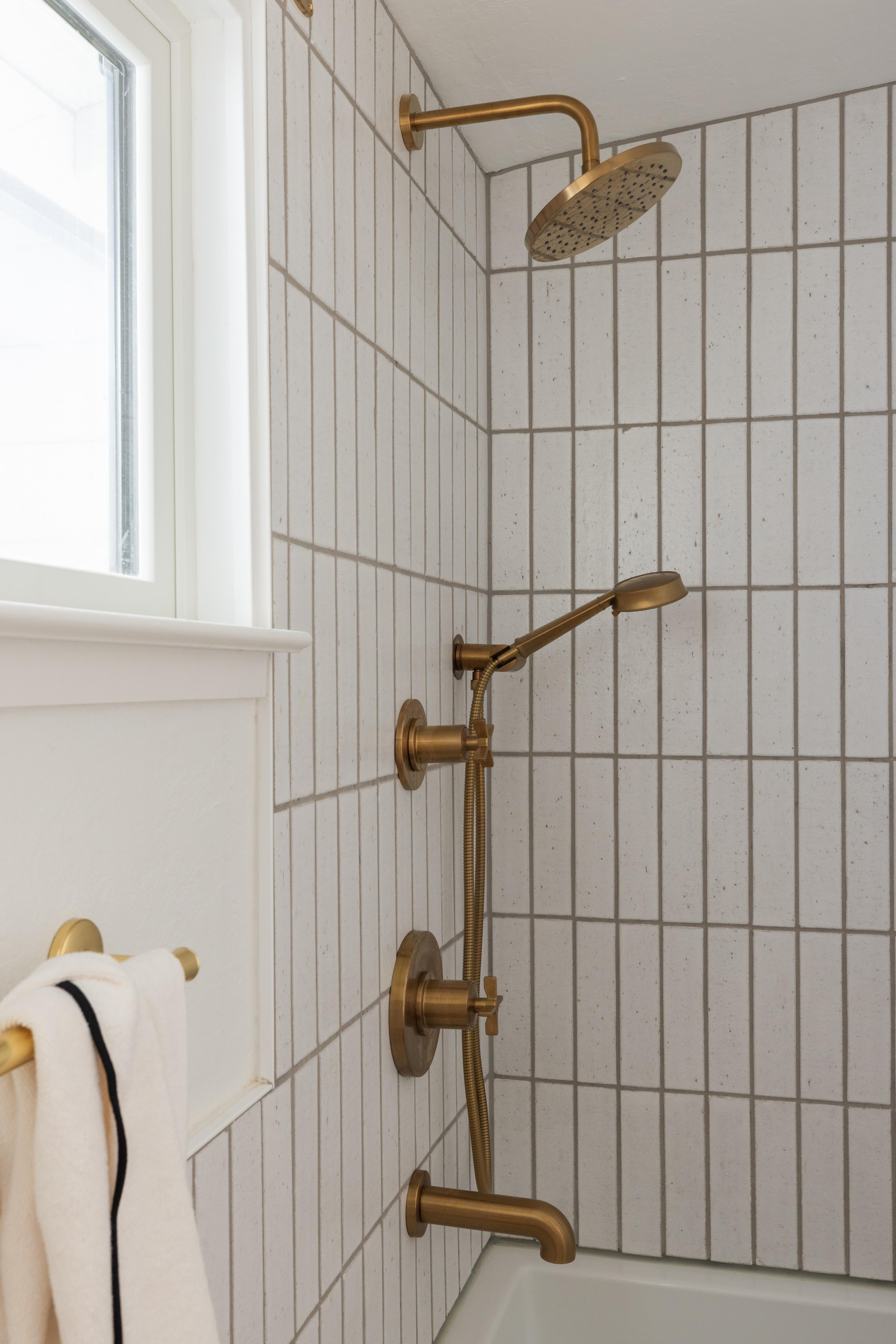 Bathroom shower with brushed brass cross-handle fixtures and rainfall head against vertical white stack tile in Carmichael home designed by Studio VAE, San Francisco interior designer.