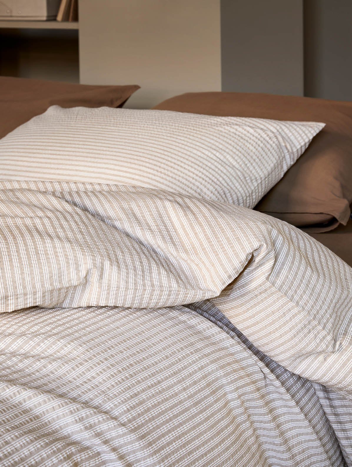 Nærbilde av en seng med hvitt kreppsengetøy med blomsterdesign. Dynen og putene har delikate, håndtegnede blomster i pastellfarger som rosa, grønn, gul og blå, som gir et friskt og sommerlig uttrykk. Sengetøyet ser mykt og behagelig ut, lagt pent over en lys seng.