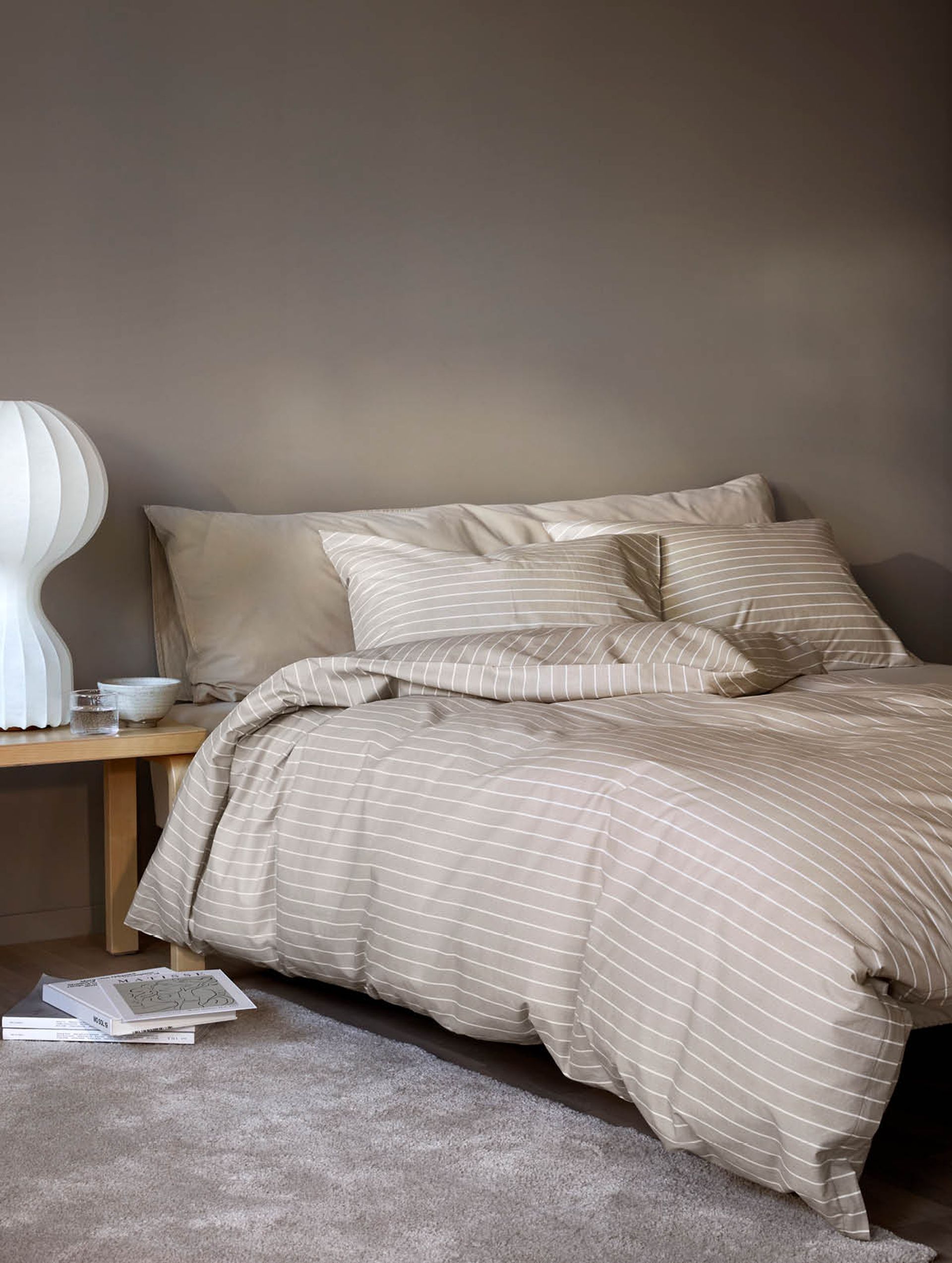 Nærbilde av oppredd seng med Høie Tor og Frøya sengesett i beige – en avslappet miks av smale striper og ensfarget bomull, som skaper en rolig og lun atmosfære på soverommet.