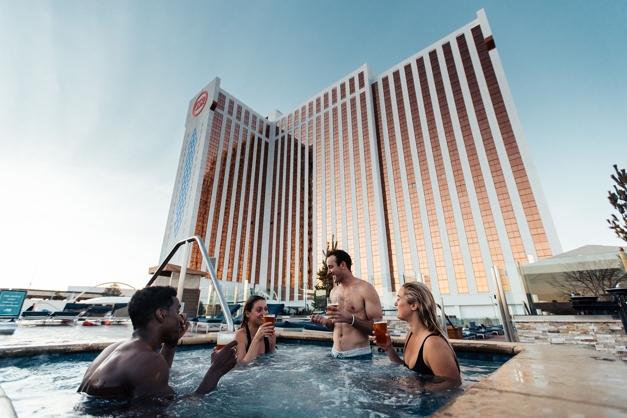 Four people are in a hot tub, with drinks in hand, in front of a large hotel building.