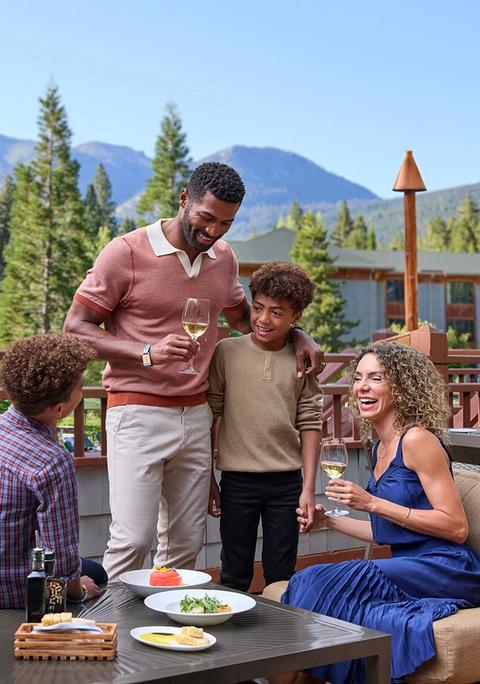 A family of three is gathered around a table on a deck, with a mountain view in the background.