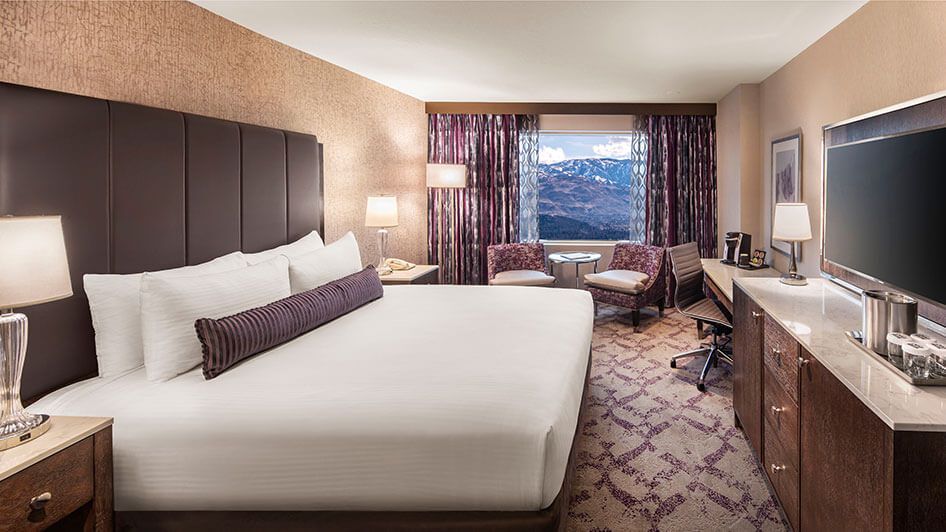 A hotel room with a large bed, two chairs, a table, and a window with a mountain view.