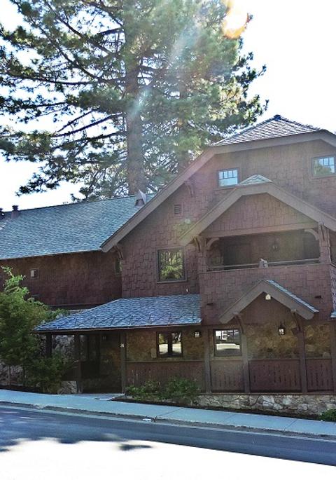 A large brown building with a gray roof and multiple windows, surrounded by trees and a road in front.