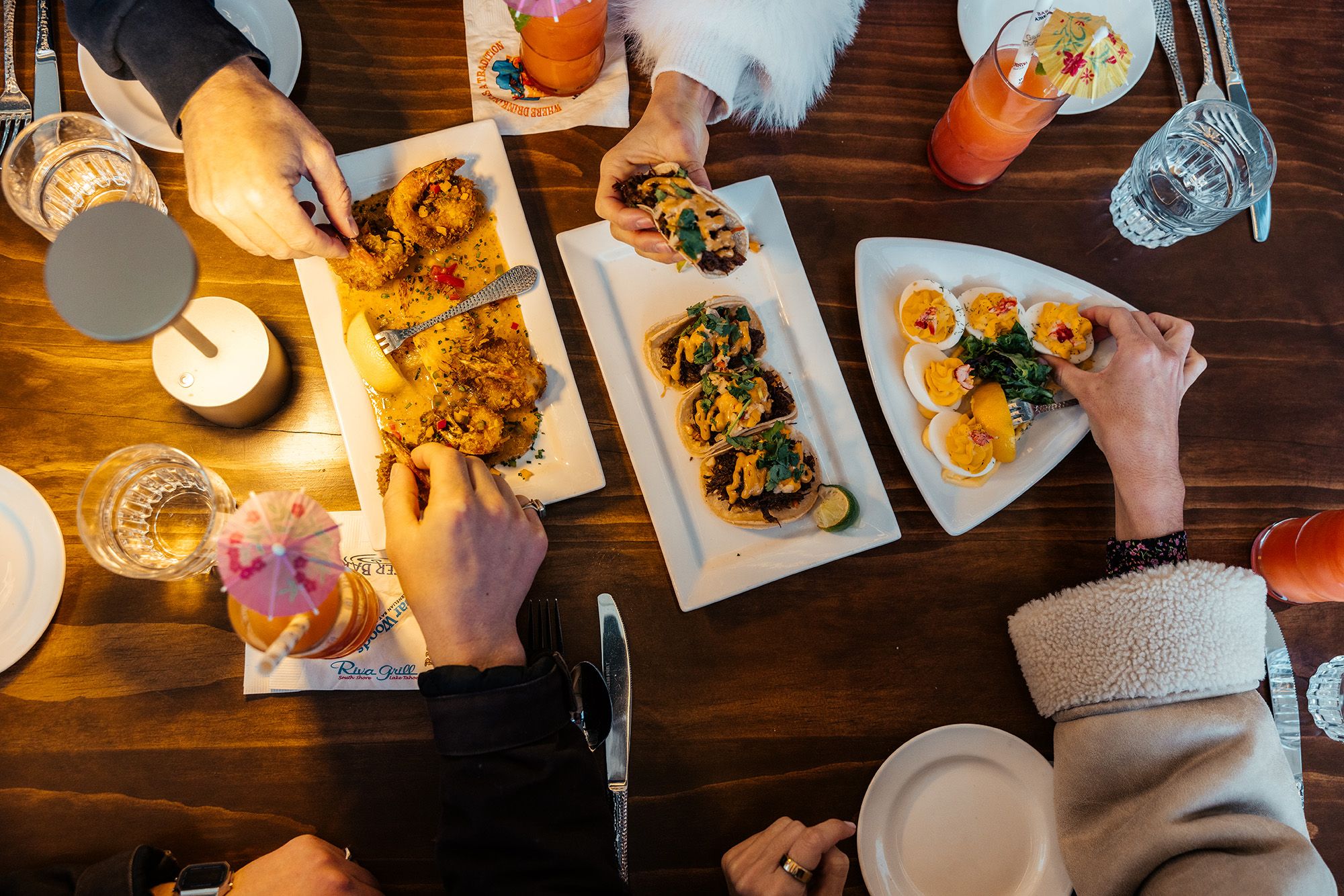 A top-down view of a table with food and drinks, with people's hands reaching for tacos and other dishes.