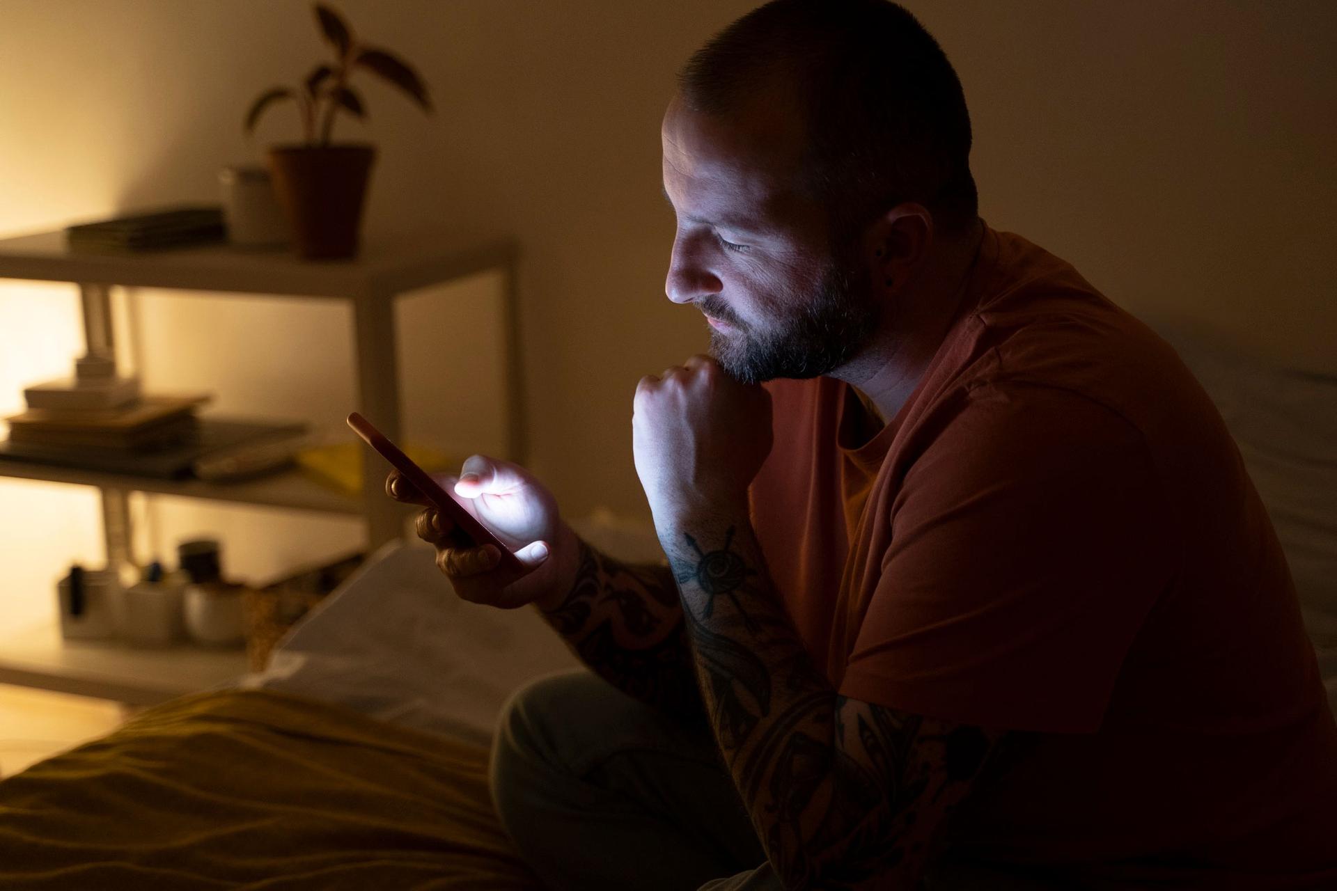 Man sitting in a dim room, looking worriedly at his phone screen, fist raised near his chin.