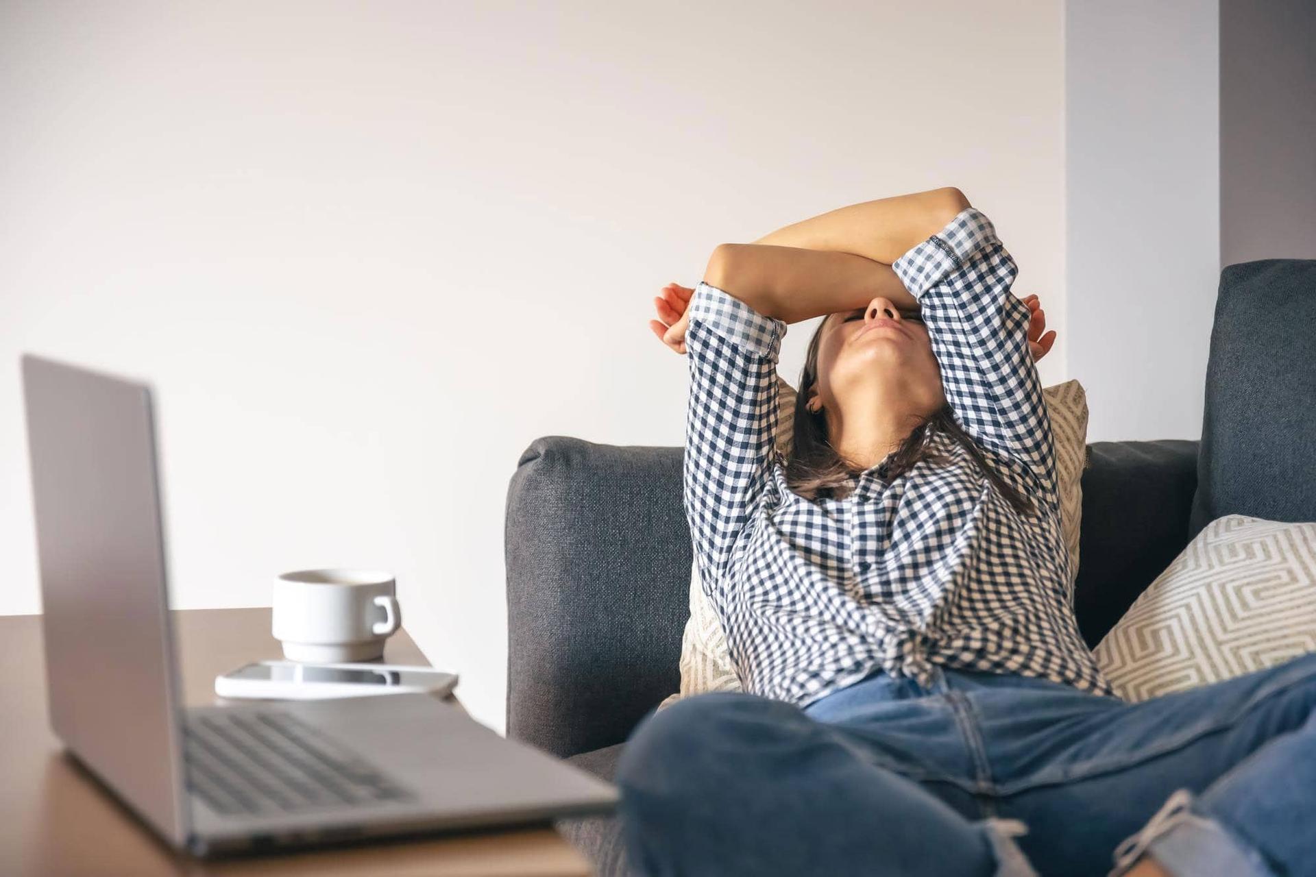 Woman sitting on a sofa with her arms crossed above her head, an open laptop and a coffee cup on the table in front of her.