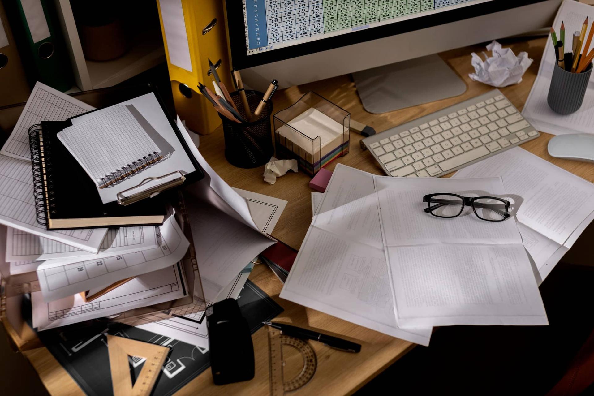 Una scrivania ingombra di fogli, cartelle e appunti, con un computer acceso sullo sfondo.