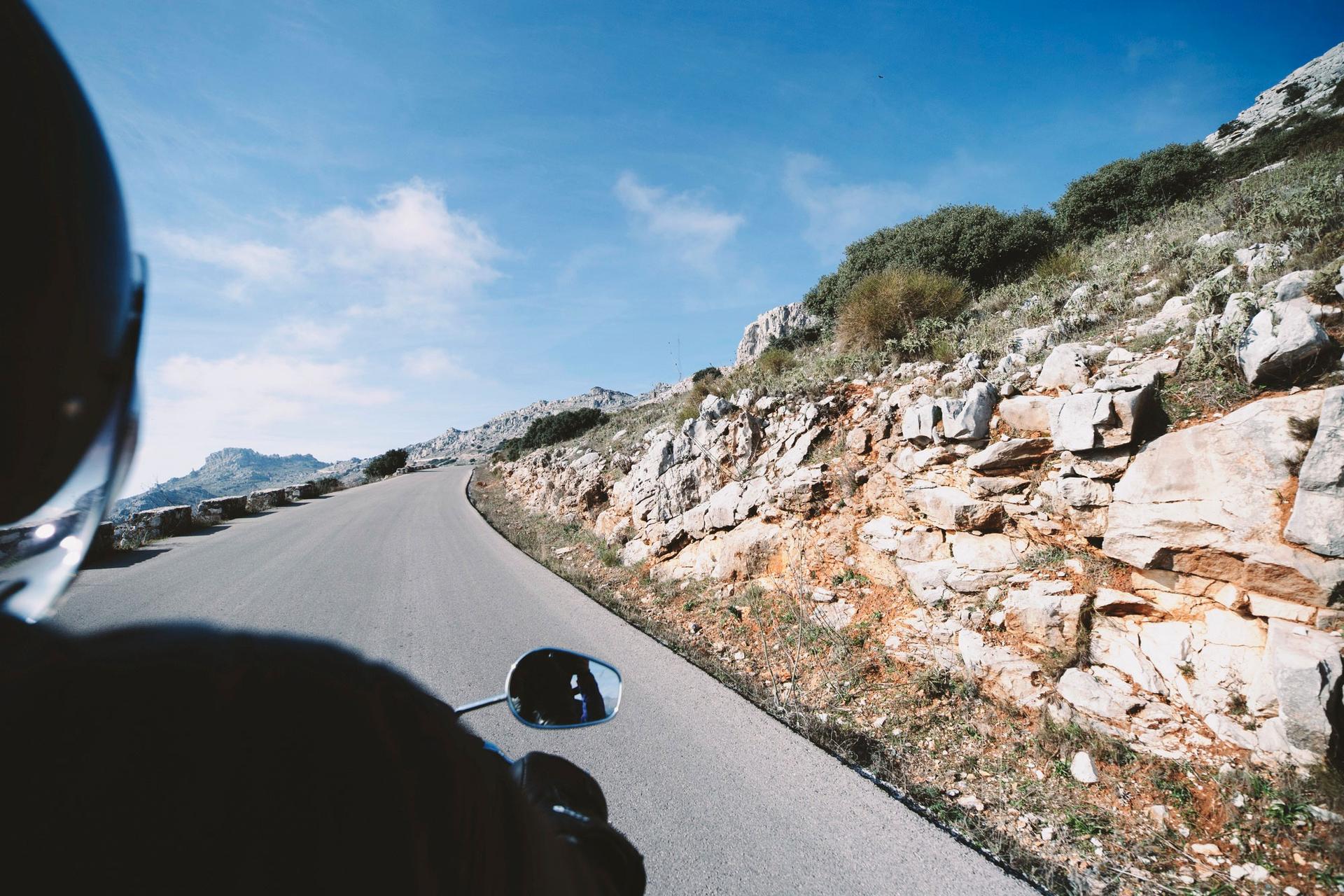 Vista da un motociclista su una strada di montagna tortuosa, tra rocce e vegetazione, sotto un cielo azzurro.