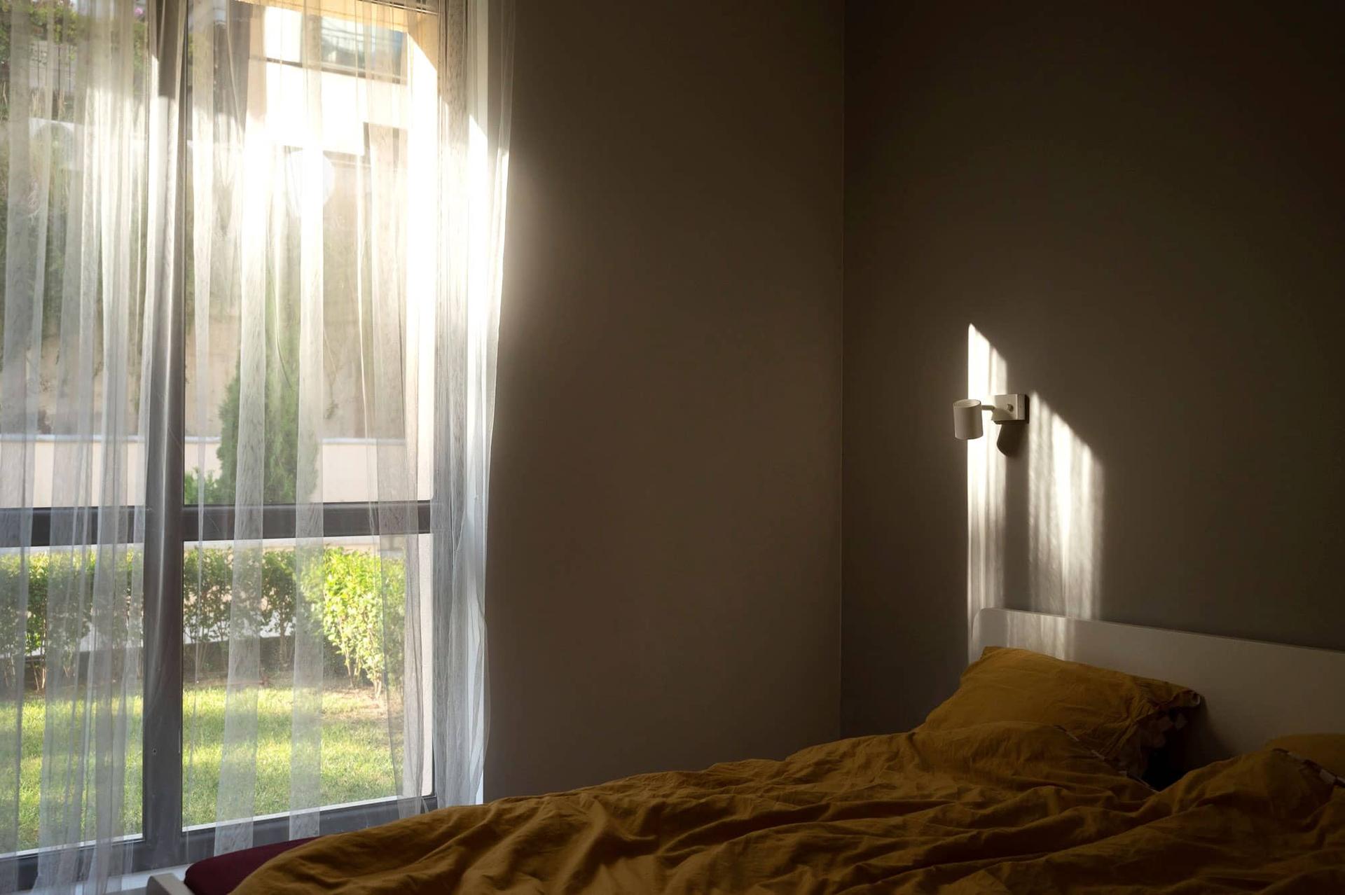 Empty bedroom with rumpled yellow bedding, sheer curtains, and morning light coming through the window.