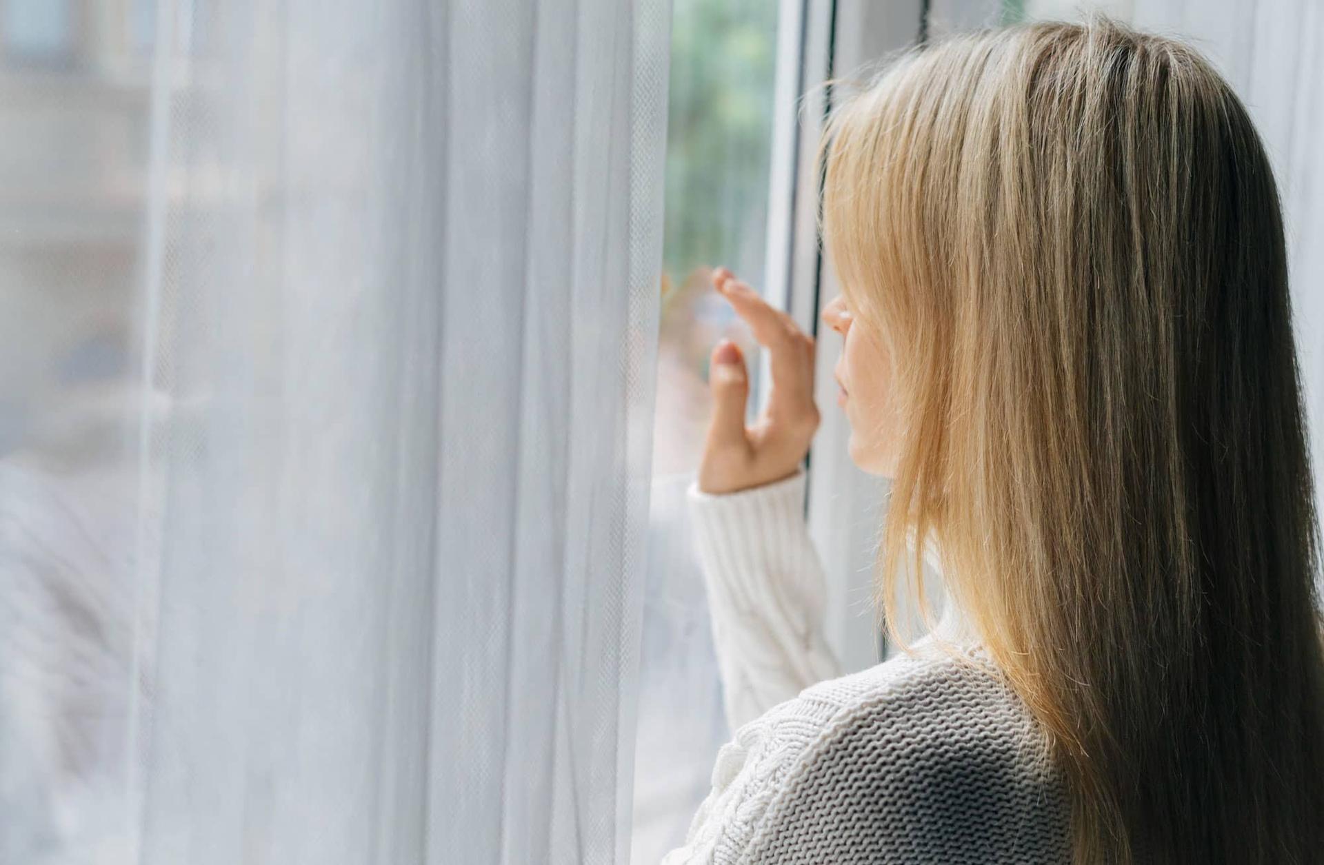 Una donna vista di spalle mentre guarda fuori da una finestra, scostando una tenda leggera.