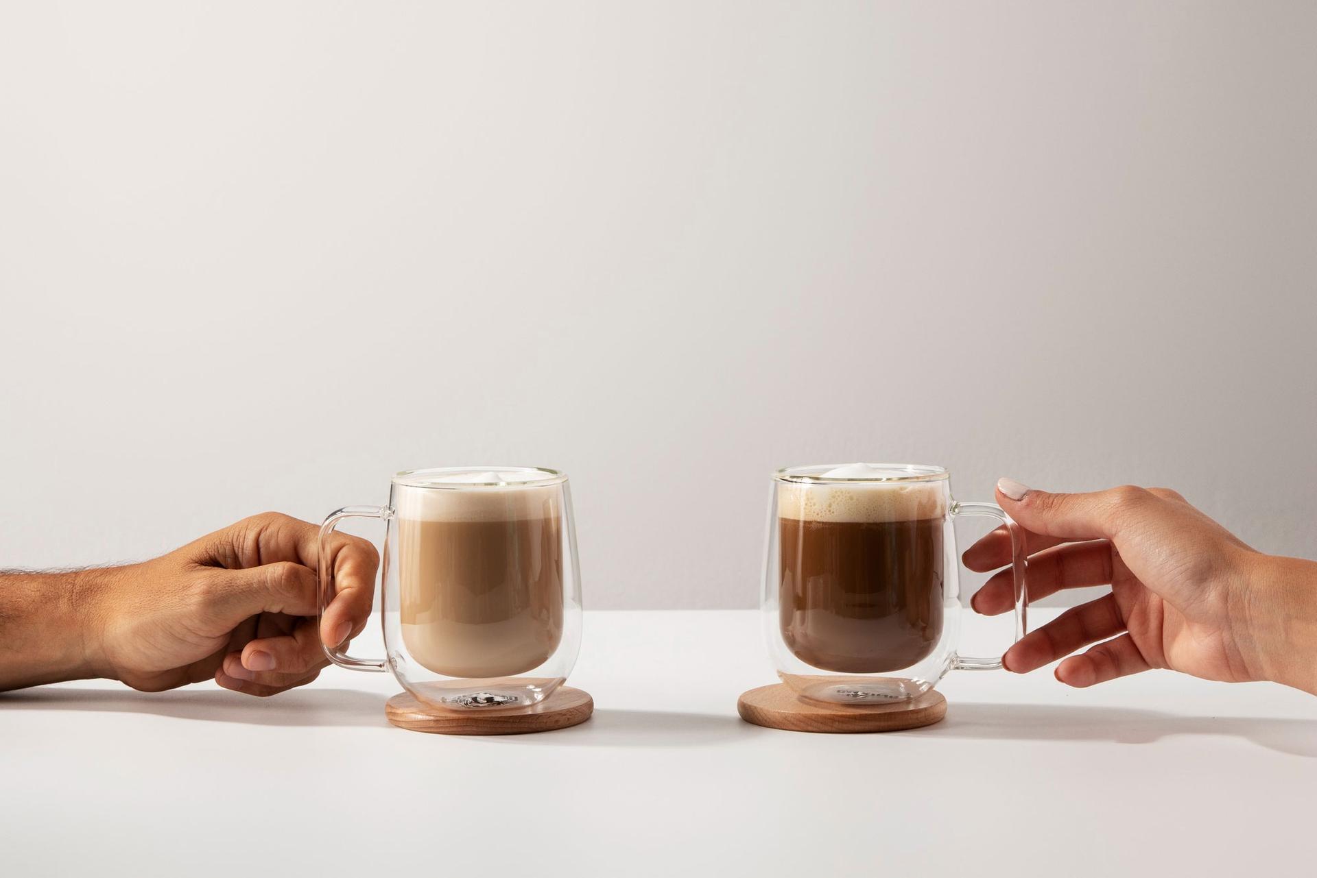 Due mani che si allungano verso altrettante tazze di vetro con caffè, su un fondo grigio chiaro.