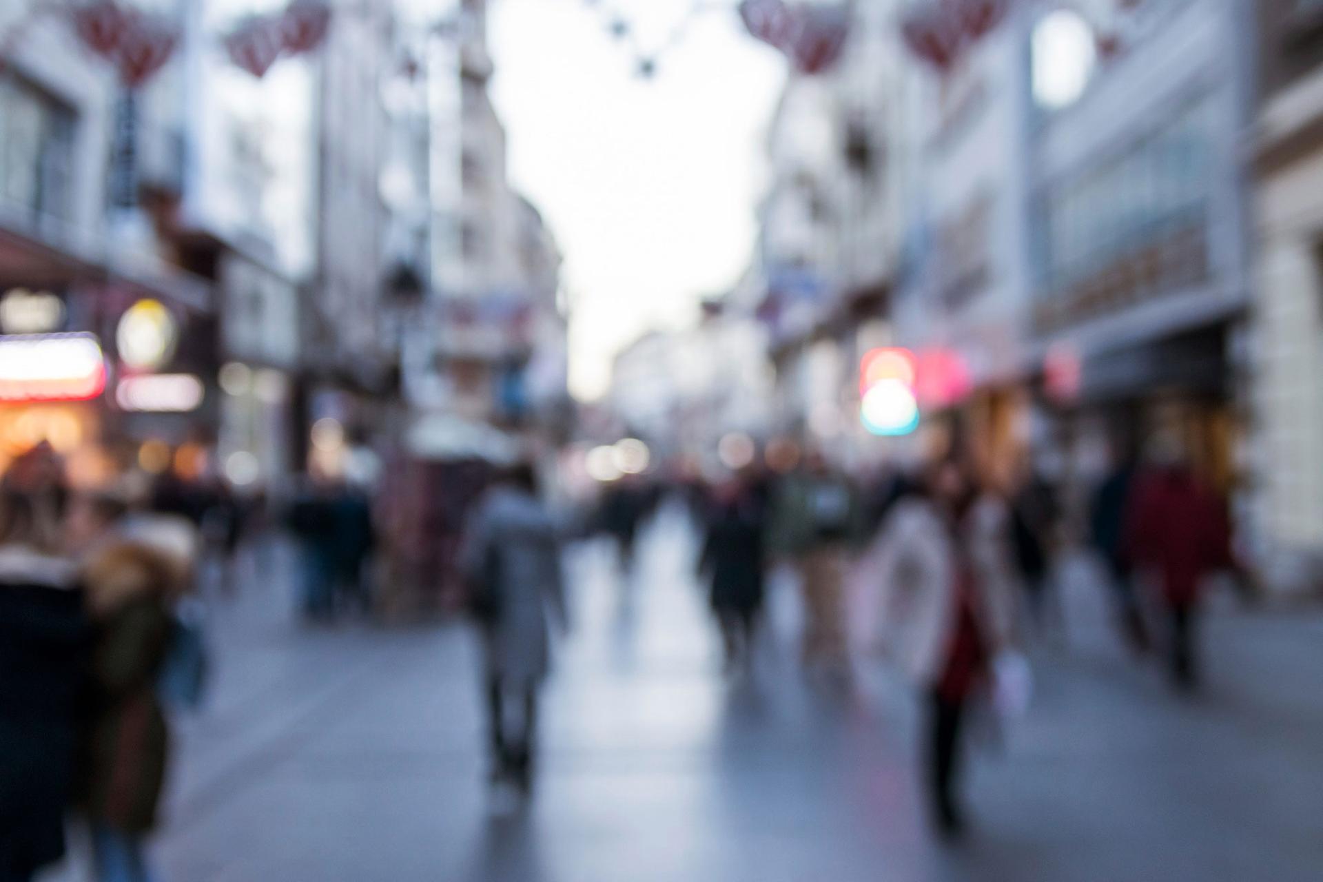 Out-of-focus crowded pedestrian street, with people walking past lit shop windows.