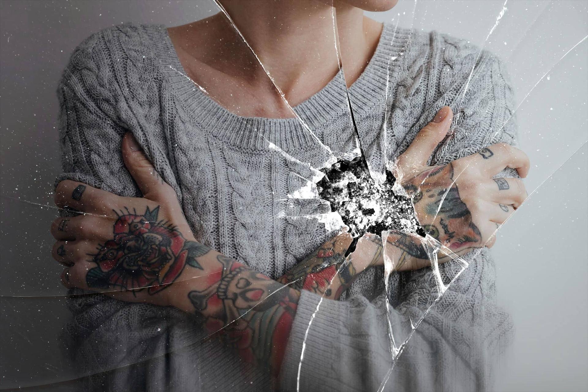 Persona con le braccia incrociate sul petto, vista attraverso una superficie di vetro incrinato.