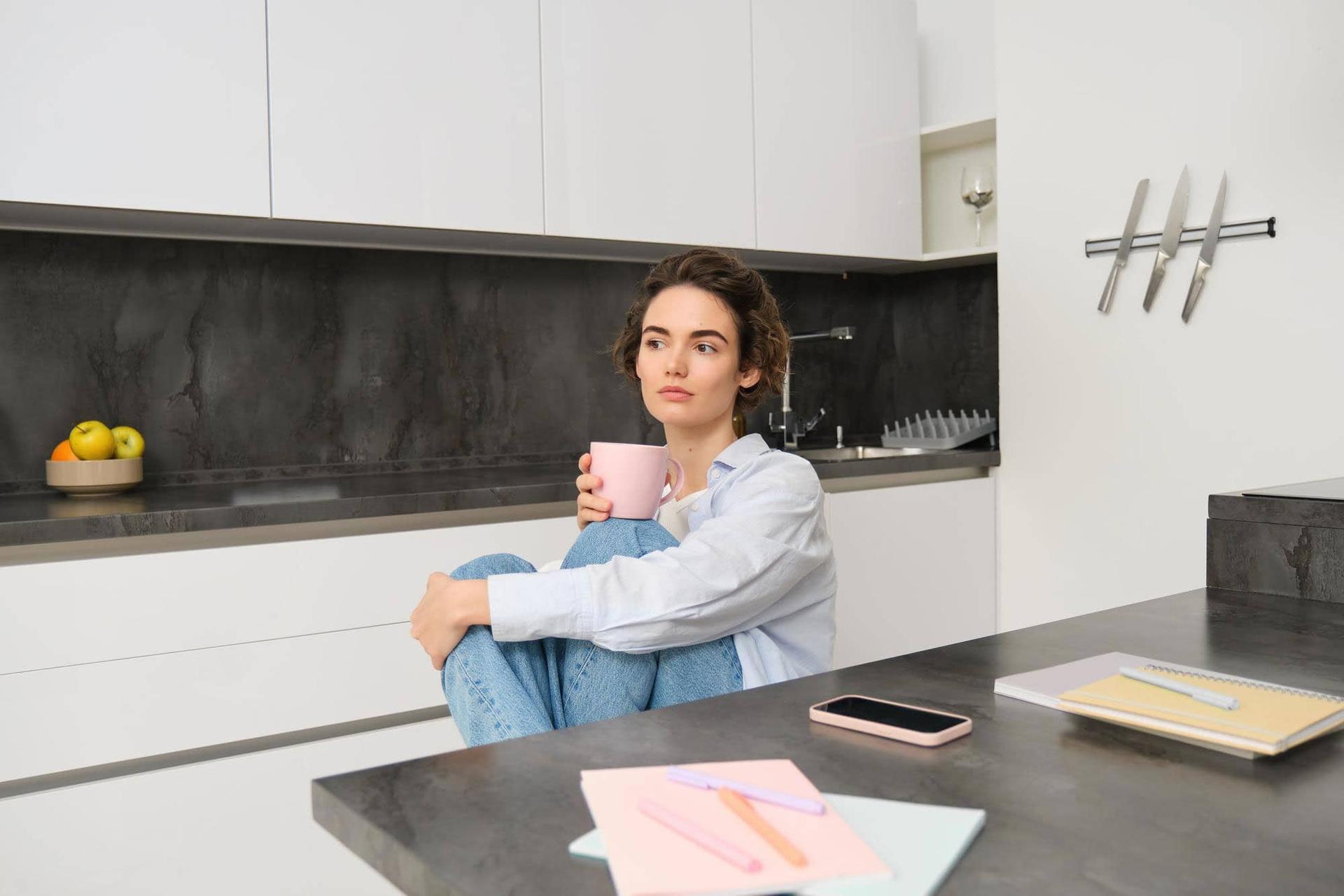 Una donna seduta su un bancone di cucina, ginocchia strette al petto, con una tazza in mano.