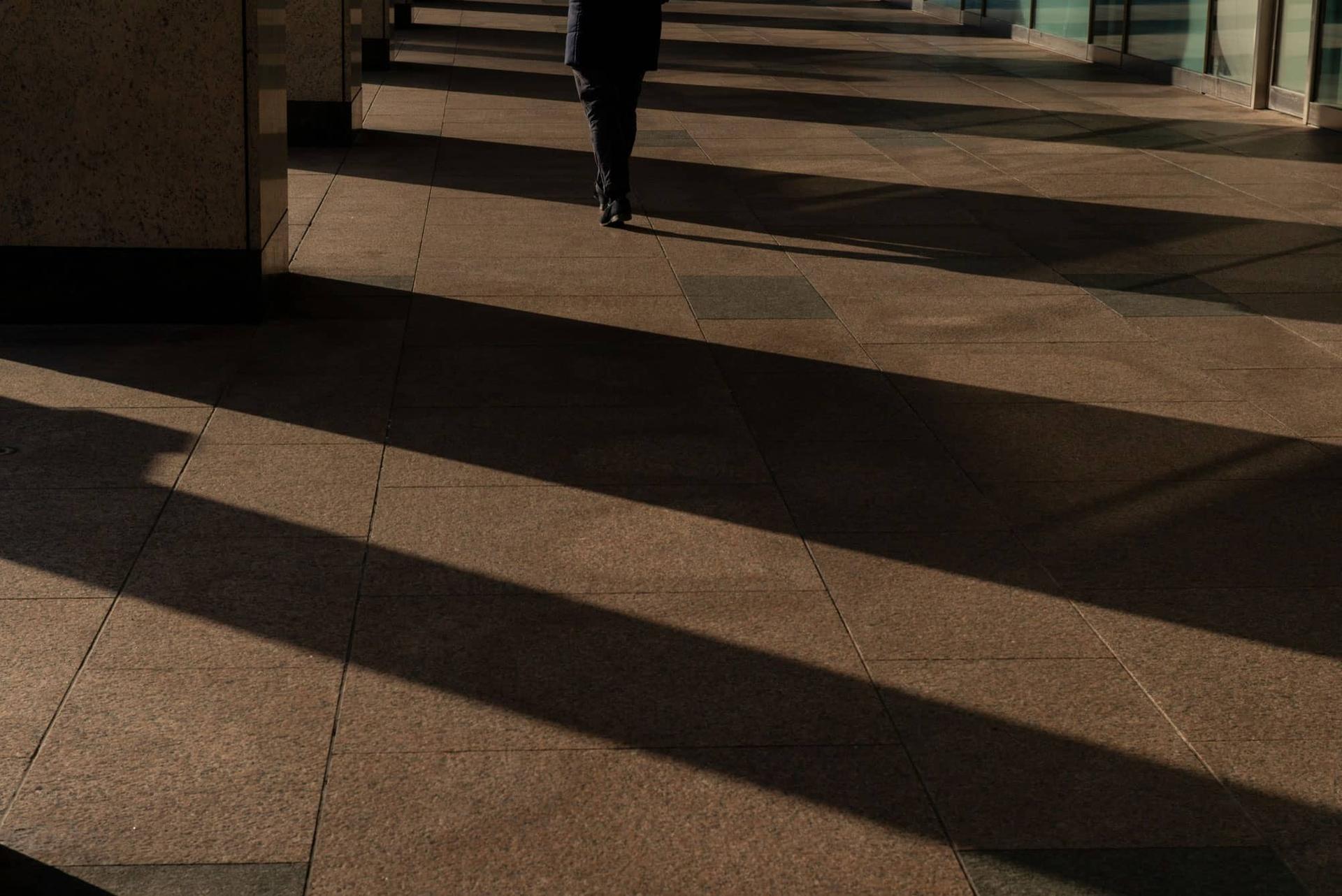 Una persona di spalle che cammina in un corridoio, con lunghe strisce d'ombra sul pavimento.