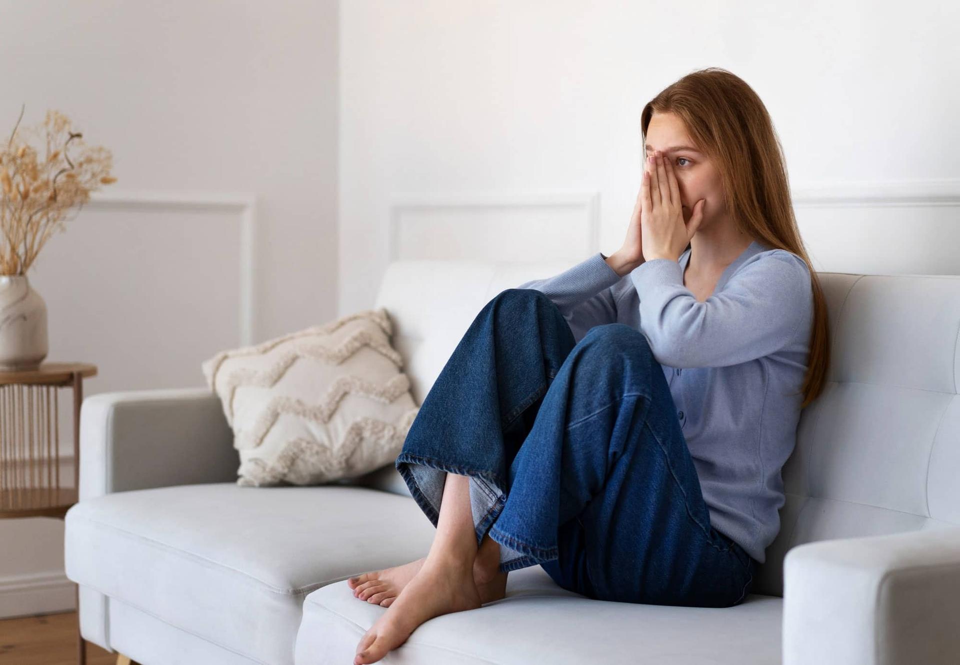 A woman sitting on a light-coloured sofa with her knees drawn up, hands partly covering her face, looking tensely to one side.