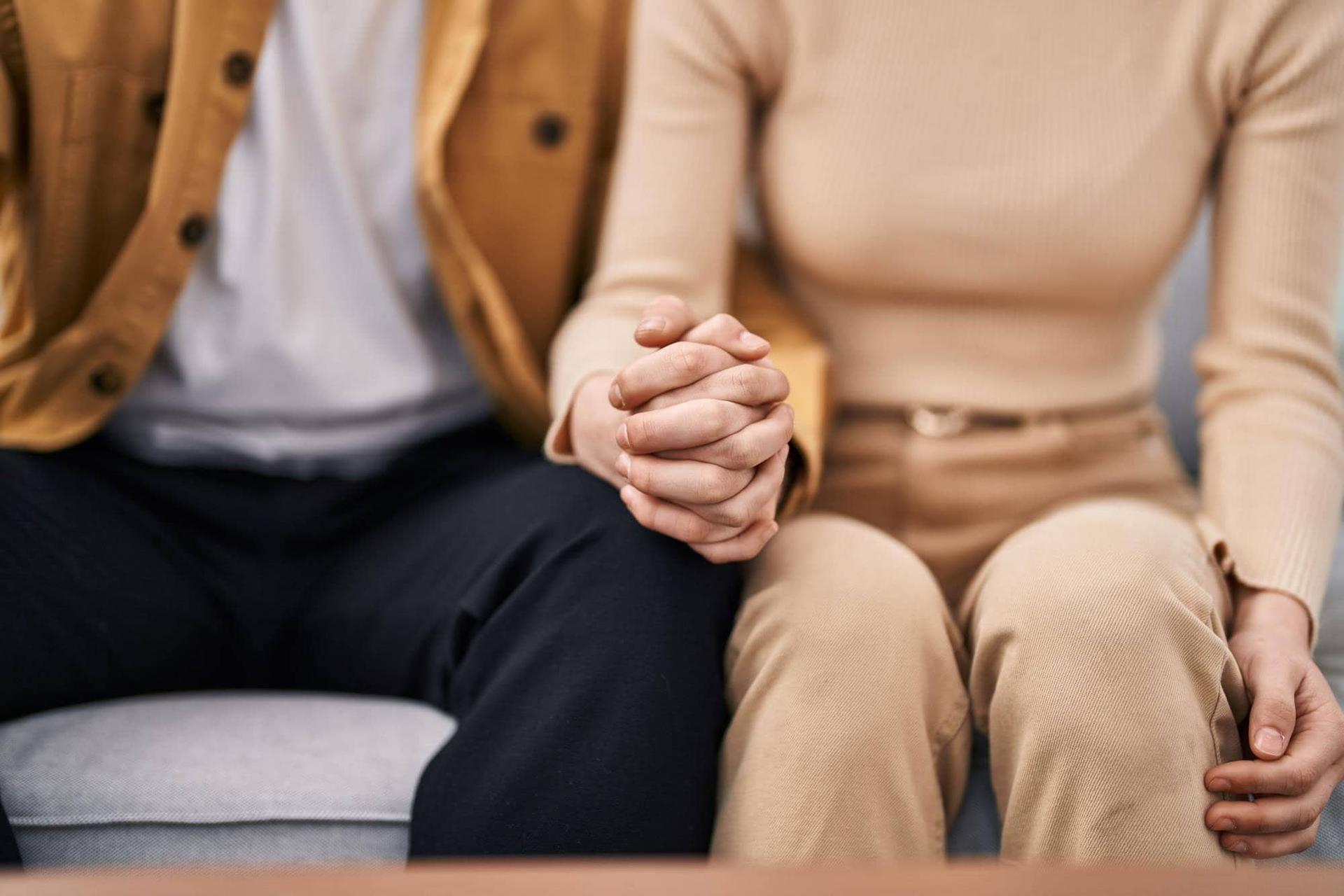Two people sitting close together, hands clasped, close-up framing on torso and knees.