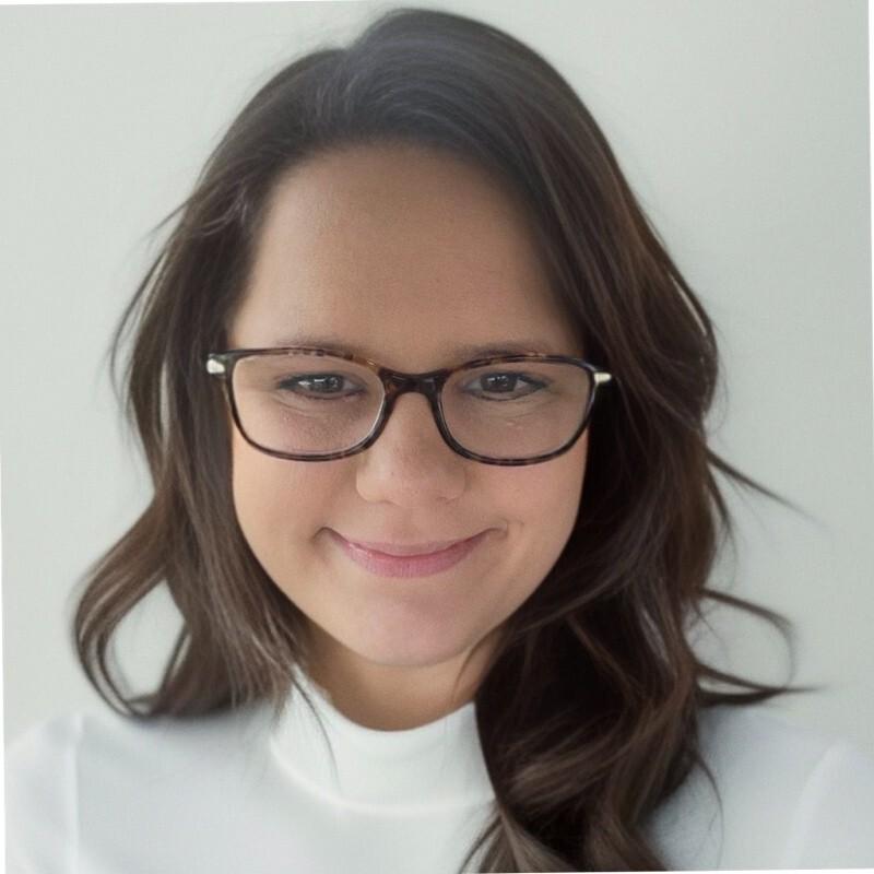 Mulher sorrindo, com cabelos castanhos e óculos, vestindo uma blusa branca. O fundo é claro e neutro.