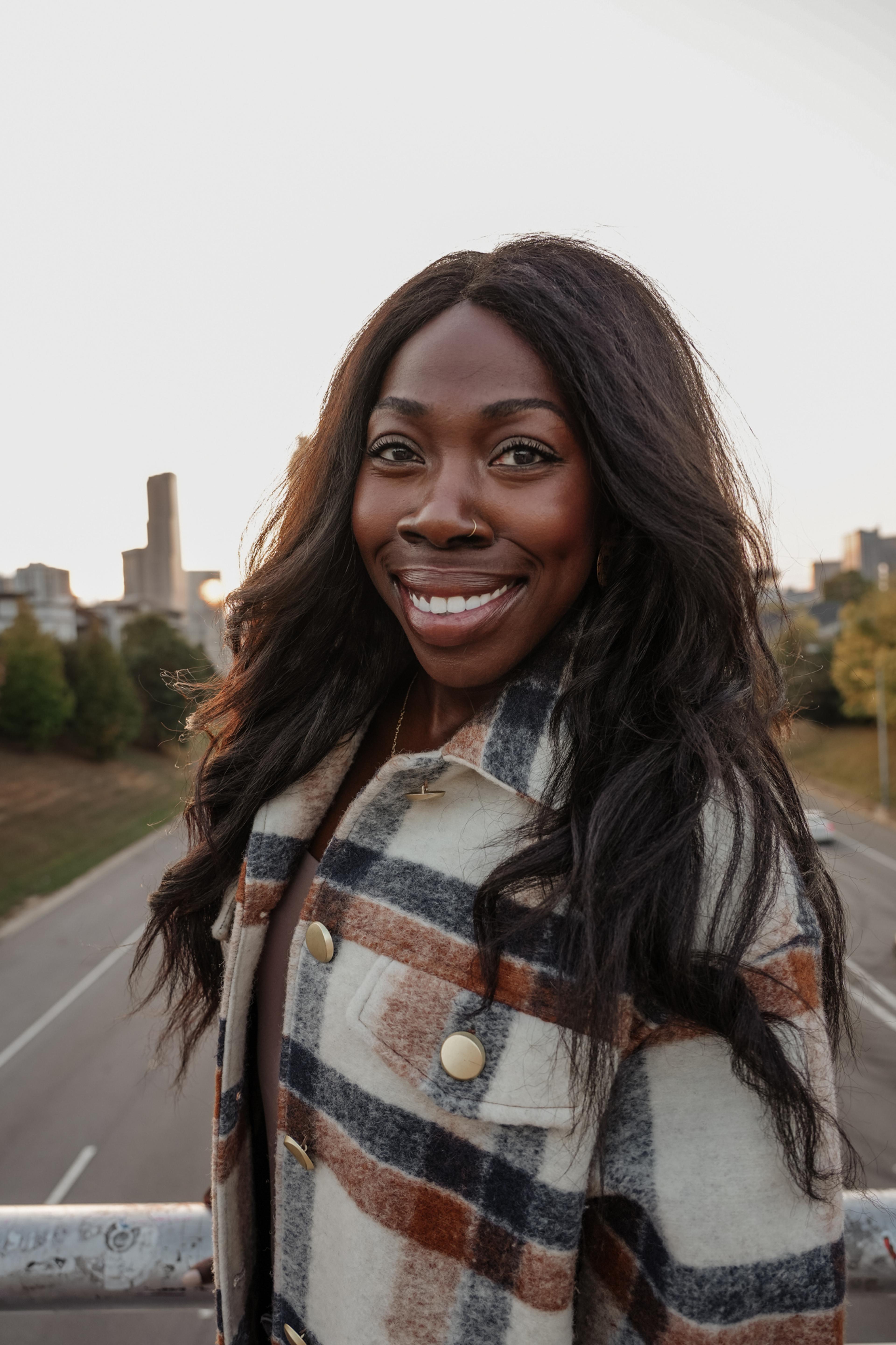 A smiling woman with long, wavy hair stands on a bridge, wearing a plaid coat. The city skyline is visible in the background during sunset.