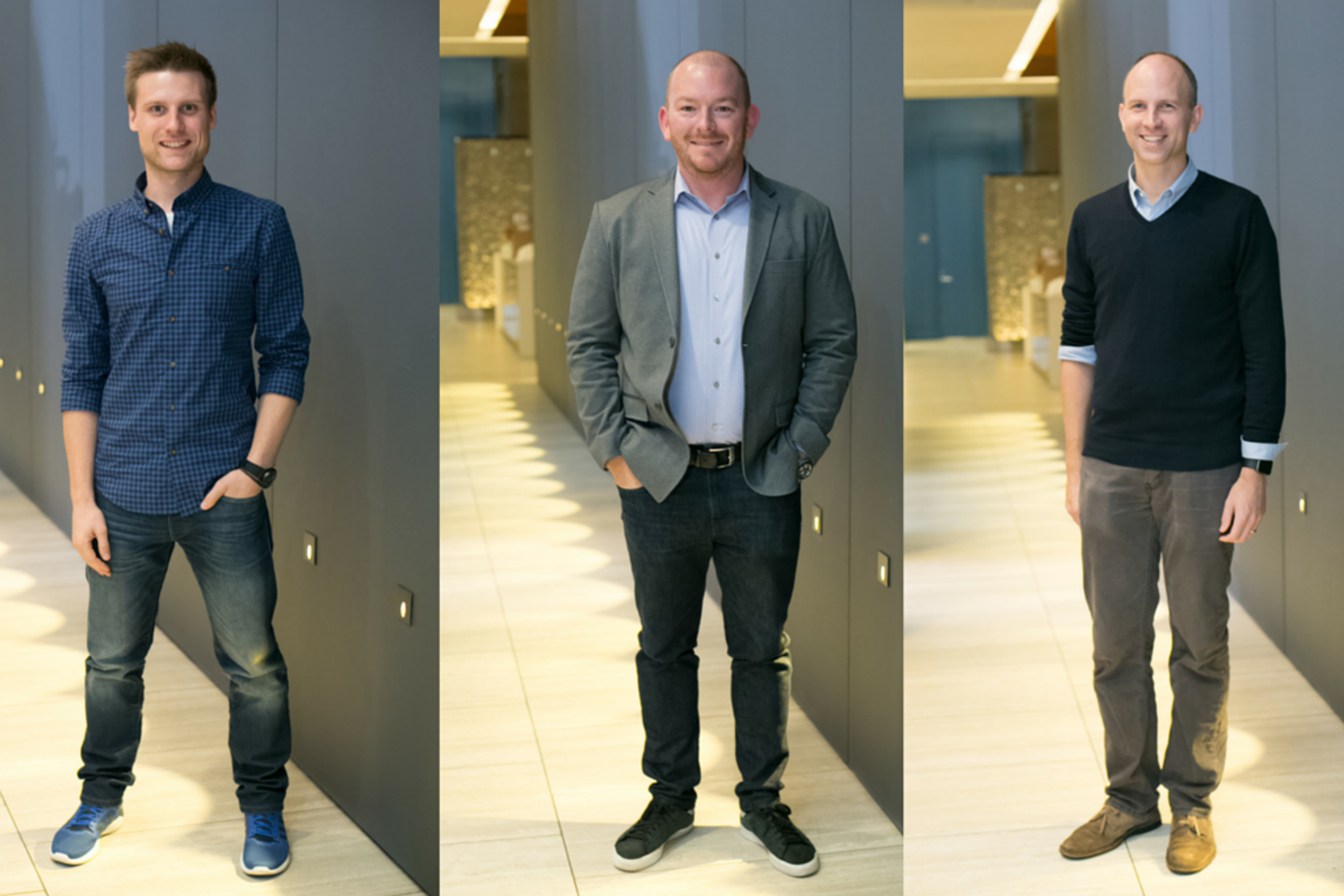 Three men stand in a modern hallway. The first man wears a blue checkered shirt, the second is in a gray blazer, and the third is dressed in a black sweater.