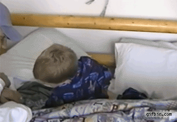 A young child is sleeping on a bed, rolling over and adjusting their position while still asleep. The bedding features a colorful pattern.