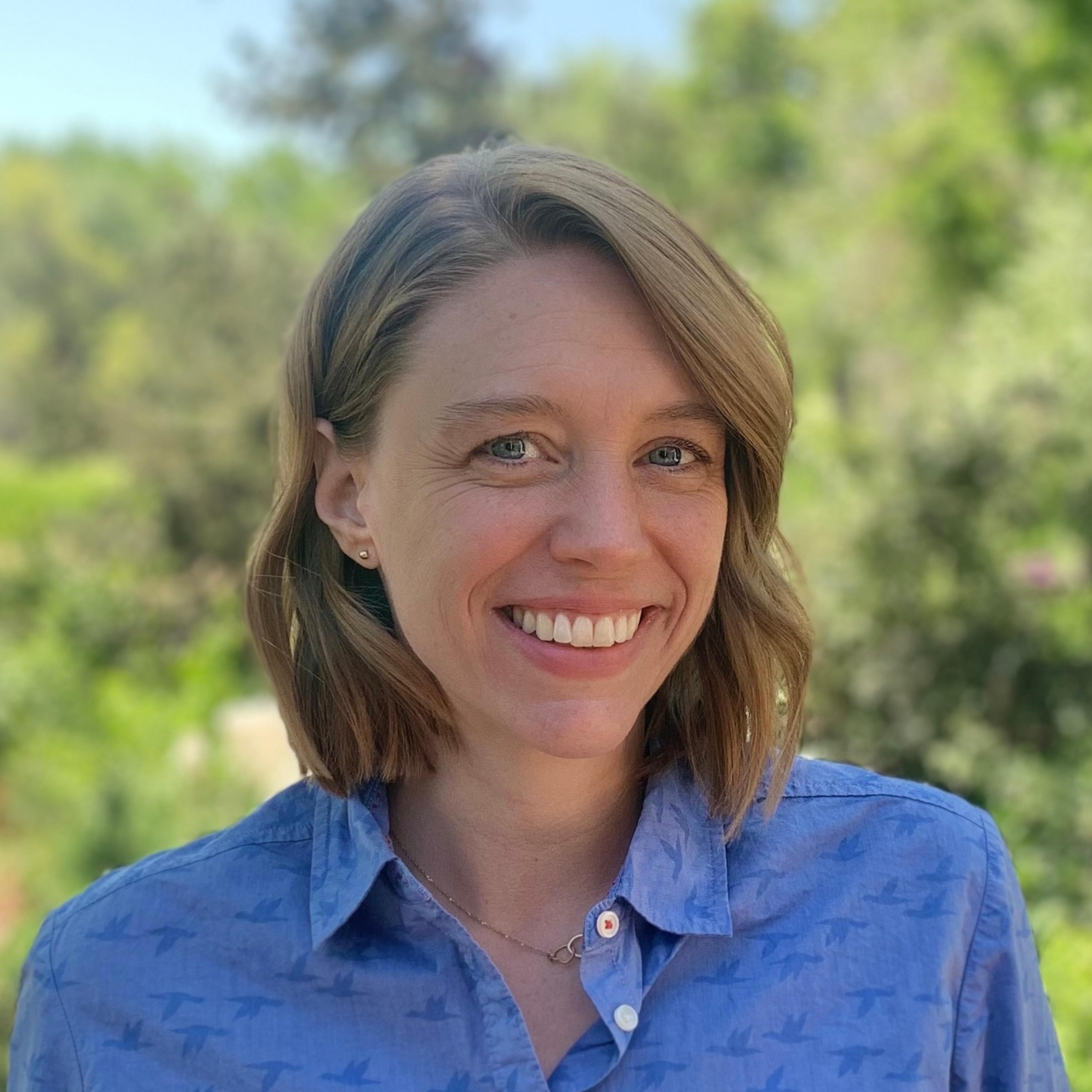 Femme souriante avec des cheveux mi-longs, portant une chemise bleue, devant un arrière-plan verdoyant.