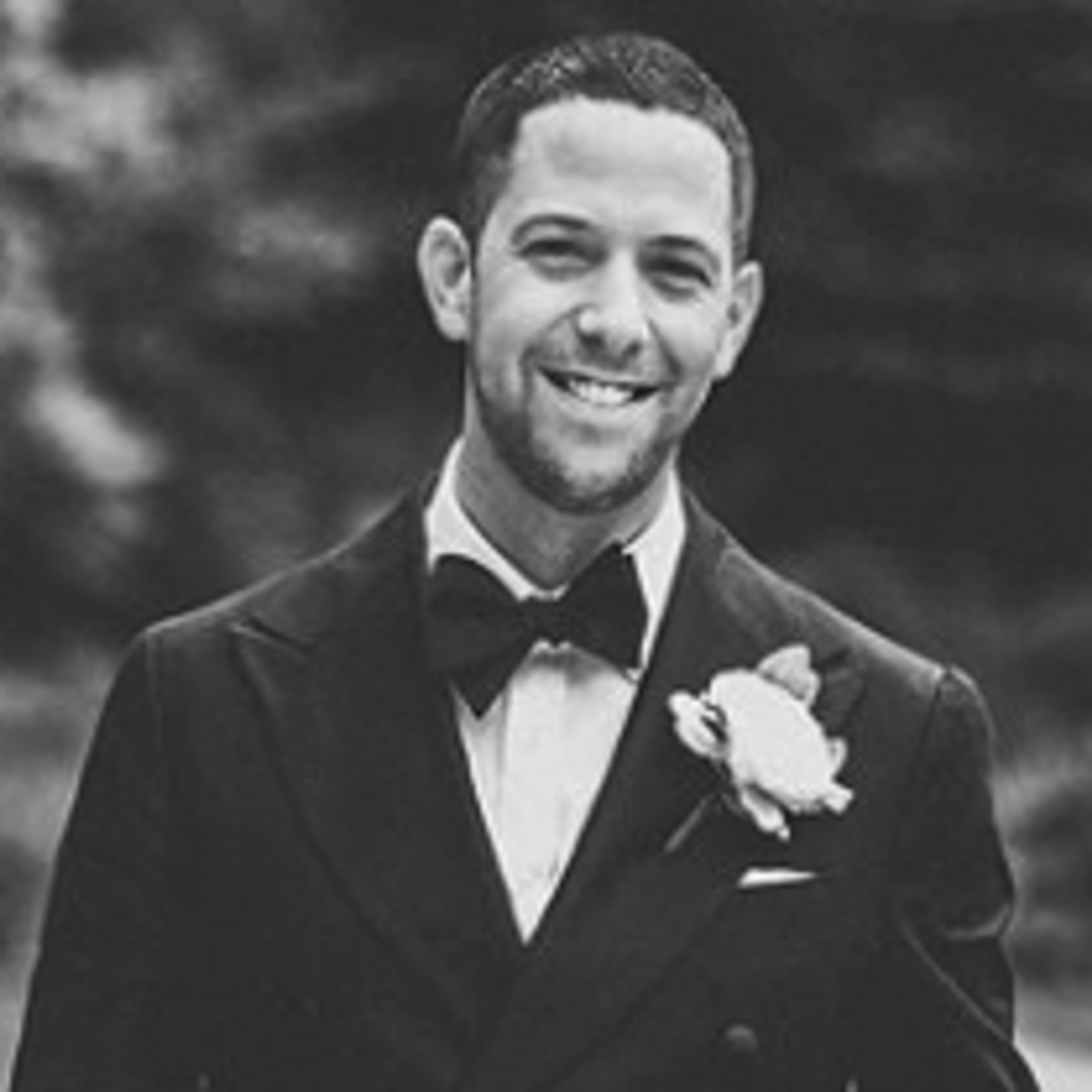 A smiling man in a formal black suit and bow tie, wearing a white flower boutonniere. The background is blurred, suggesting an outdoor setting.