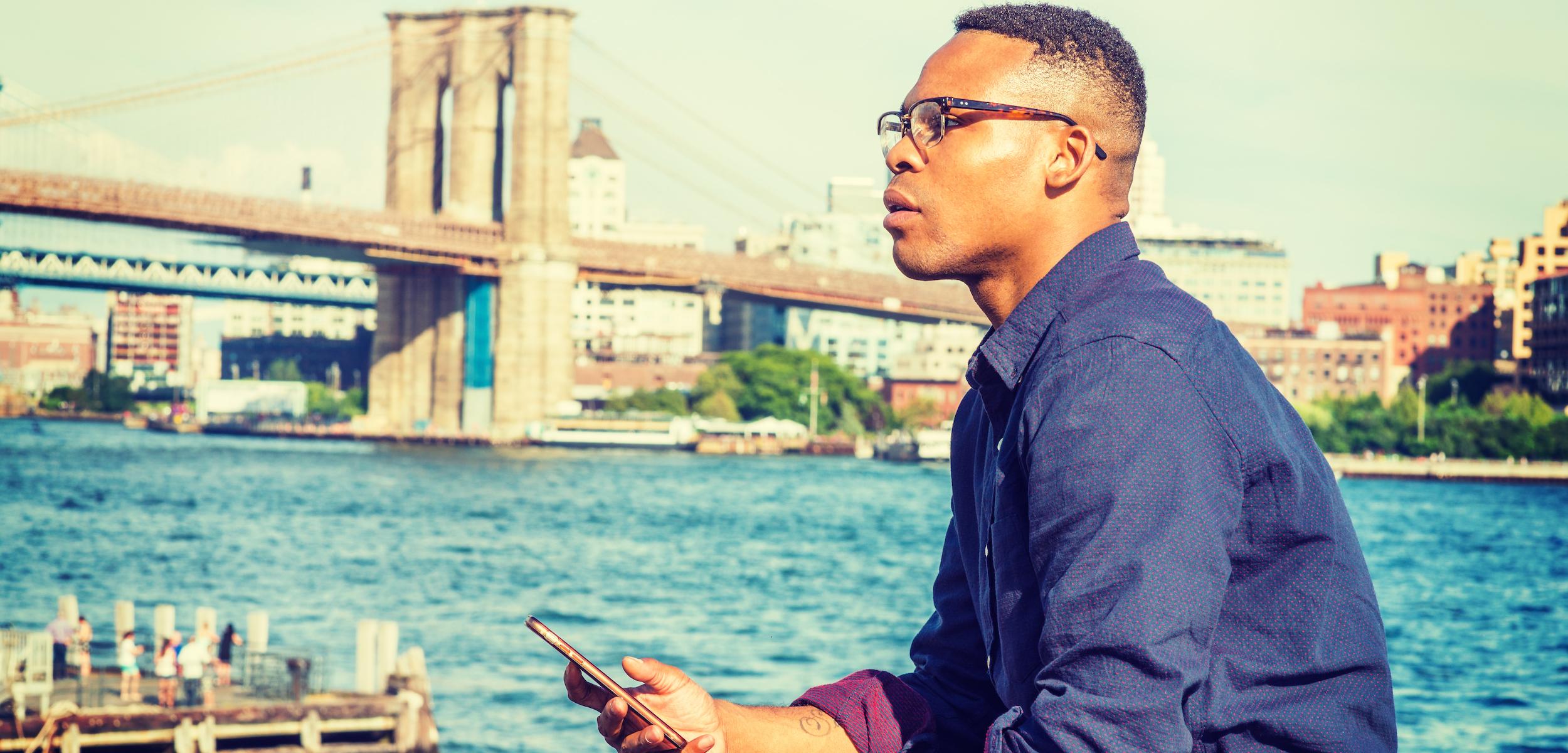 A man sits by the waterfront, looking thoughtfully at his phone with the Brooklyn Bridge in the background. The scene captures a sunny day with city buildings lining the shore.