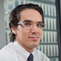 A man with glasses and dark hair is smiling at the camera, wearing a white shirt and a tie. The background features a modern building with large windows.