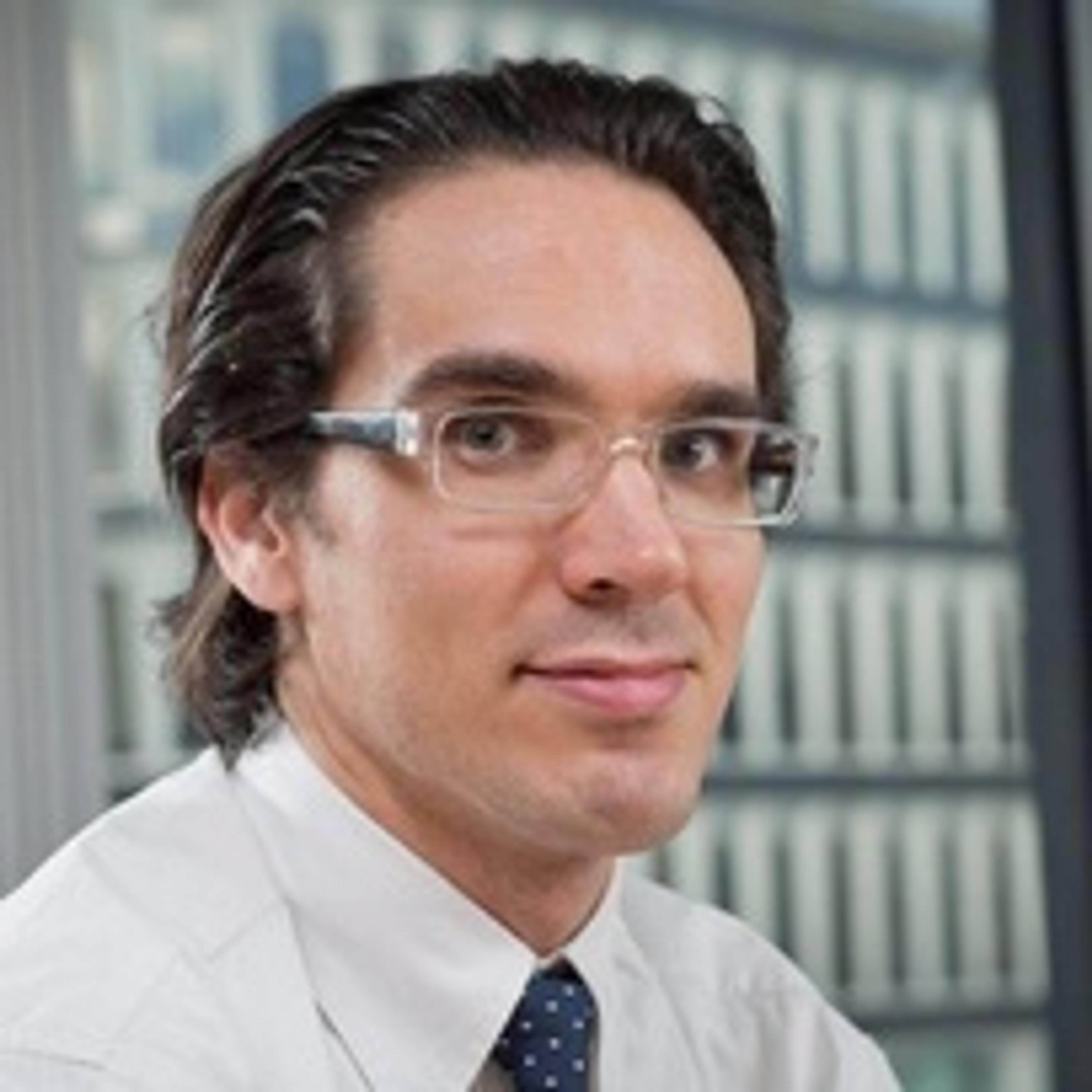 A man with glasses and dark hair is smiling at the camera, wearing a white shirt and a tie. The background features a modern building with large windows.