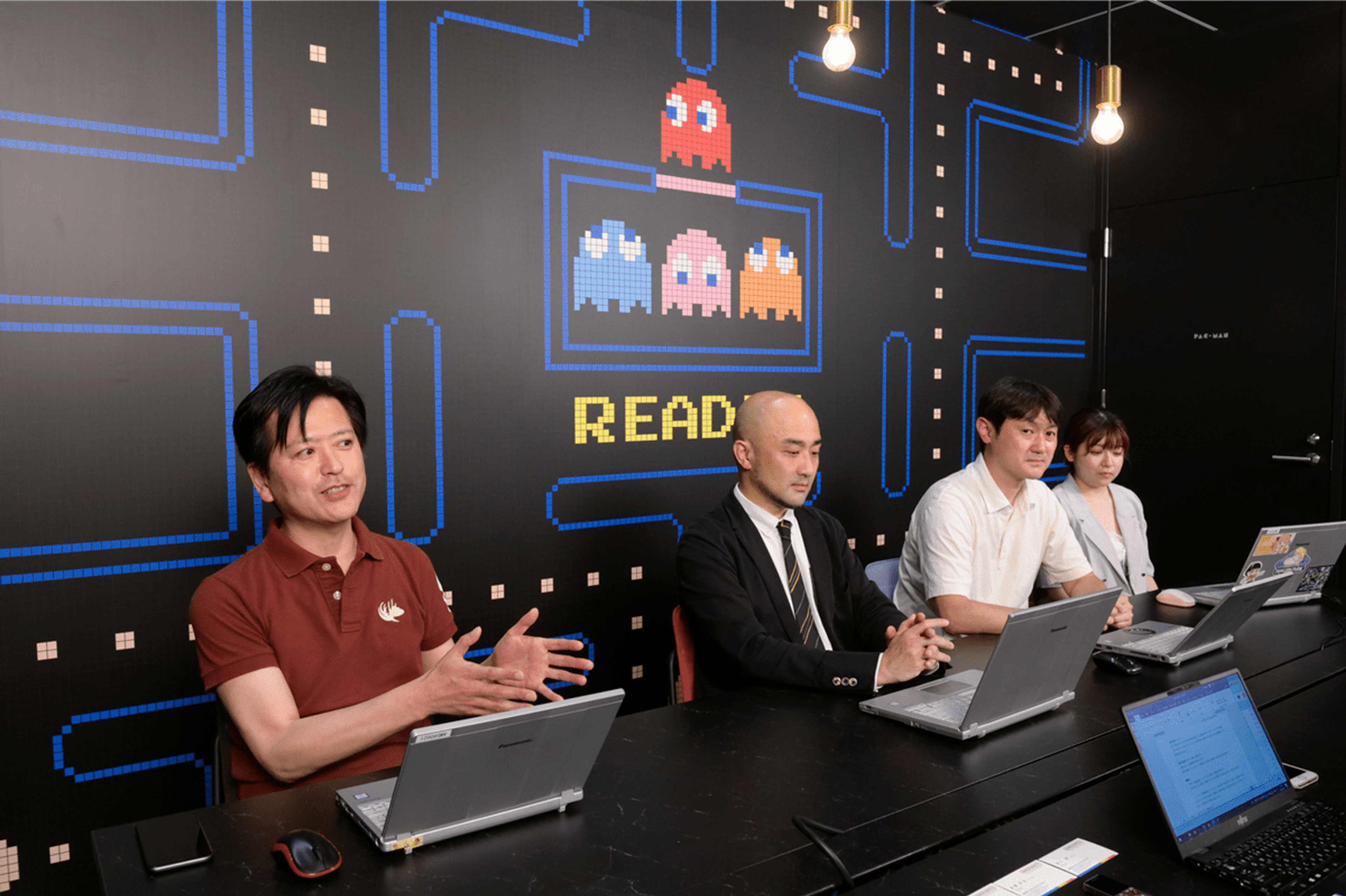 社員が四人、インベーダーゲーム背景の前に座っている。（左より株式会社バンダイナムコアミューズメント DX部 兼 マーケティング部 スペシャリスト 松本圭司氏、DX部 デジタルビジネス推進課 アシスタントマネージャー 高原 伸幸氏、DX部 OMO推進2課 マネージャー 髙橋 幸治氏、DX部 OMO推進2課 清川 舞氏）
