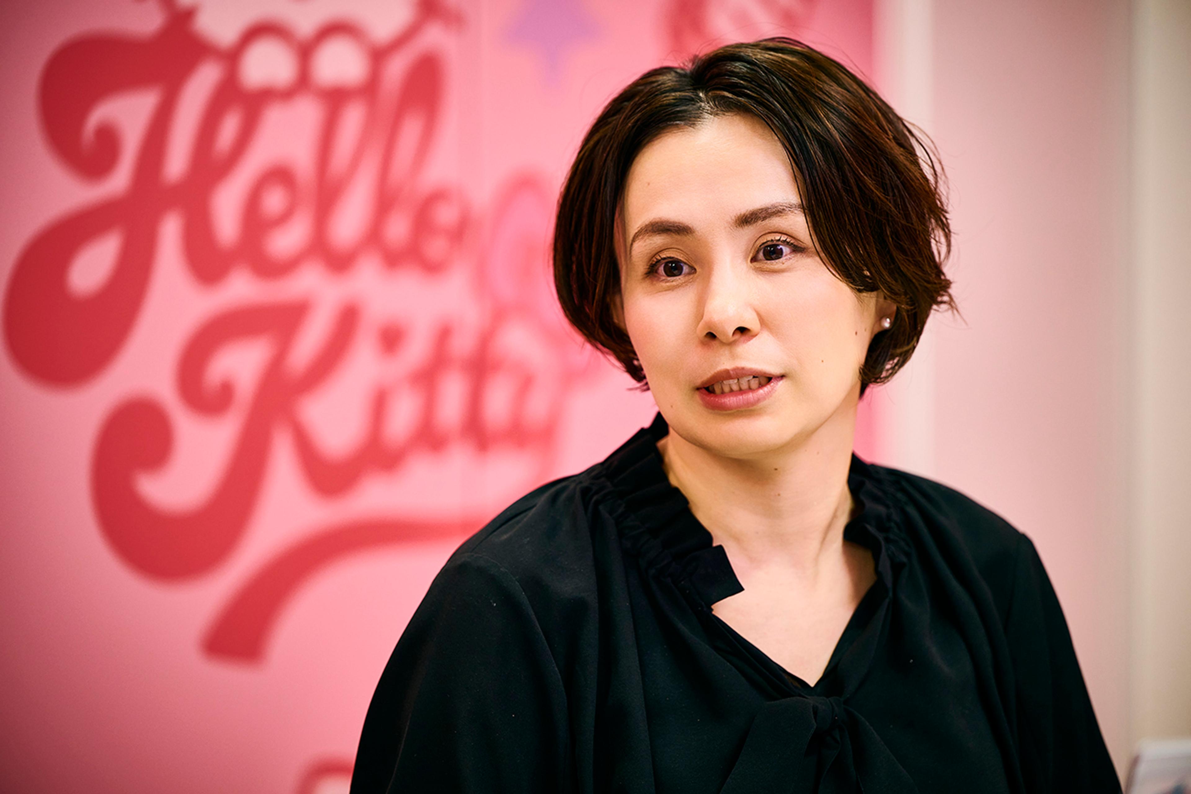 ピンクの壁を背に、ぼやけたハローキティのロゴを見ながら話す女性(サンリオ　デジタル事業本部 デジタル事業開発部 CX推進課 シニアマネージャー　鈴木 理恵氏)。