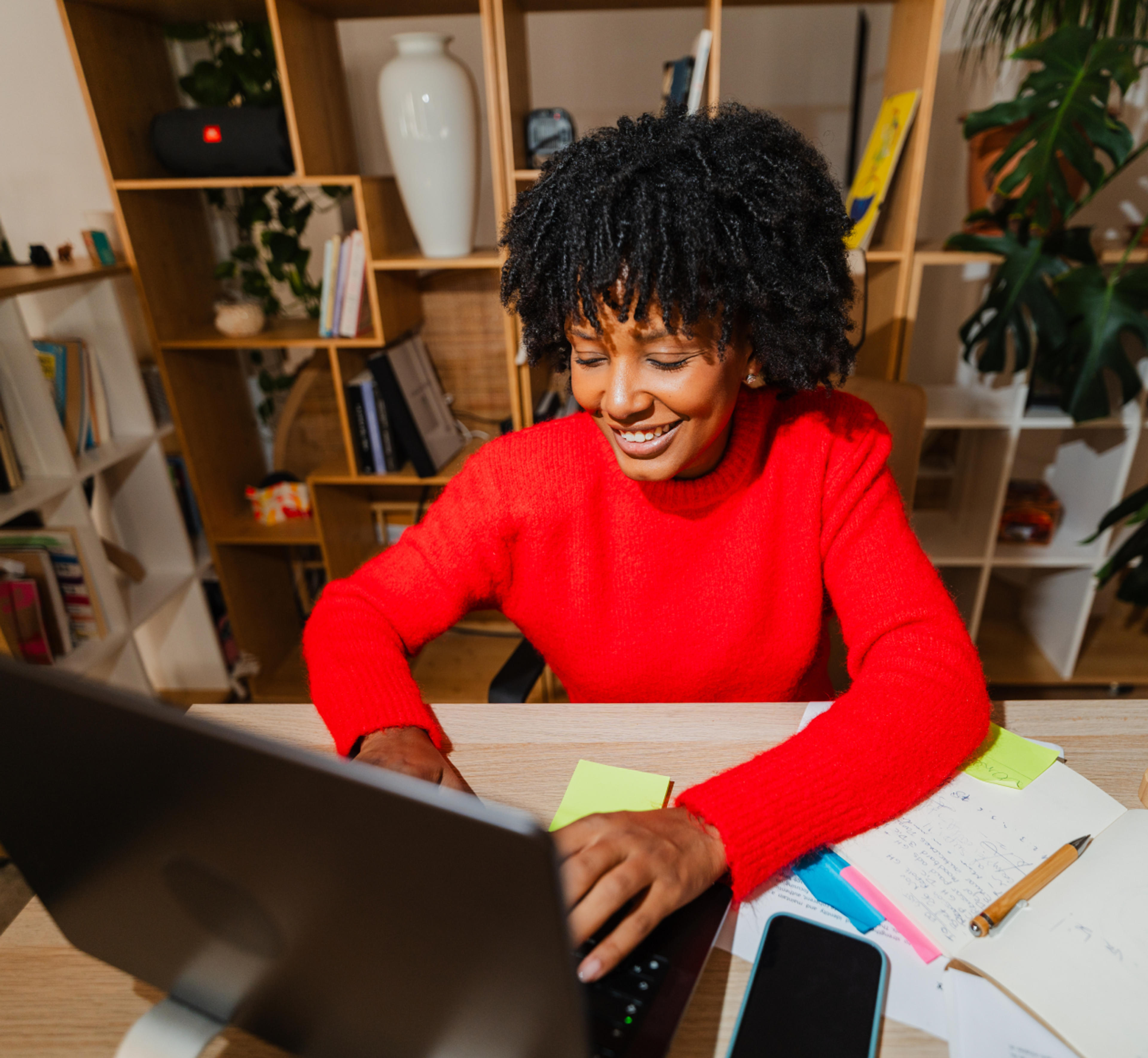 Smiling person in a bright red sweater works at a laptop in a home office, surrounded by notes, a notebook, and a phone on the desk.